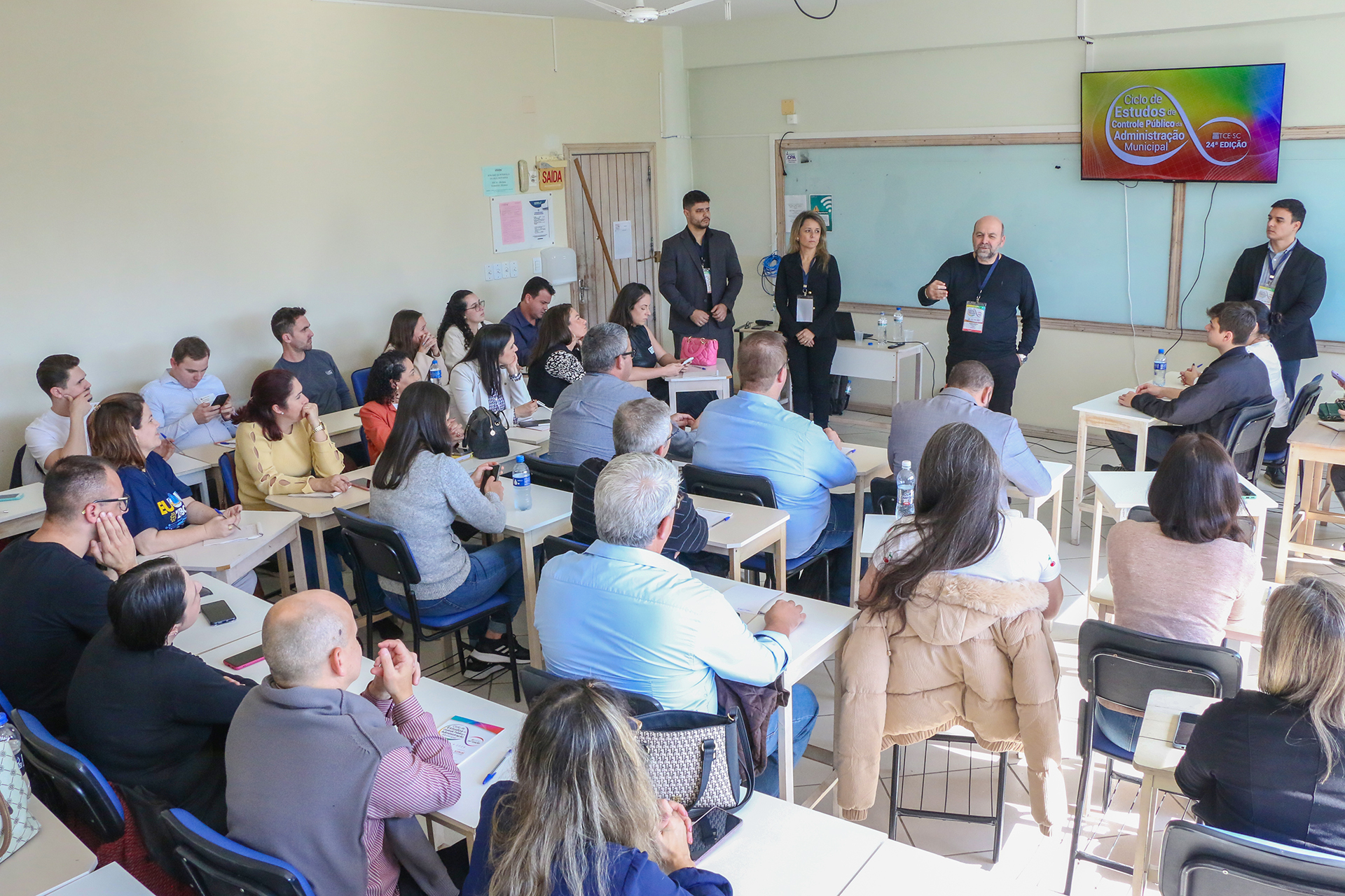 Foto de uma das oficinas do Ciclo de Estudos. Há vários participantes sentados em fileiras, em uma sala de aula. À frente, quatro membros do TCE/SC que coordenam as atividades. Entre eles, o diretor do Icon, Moisés Hoegenn, que está em pé, falando ao público.