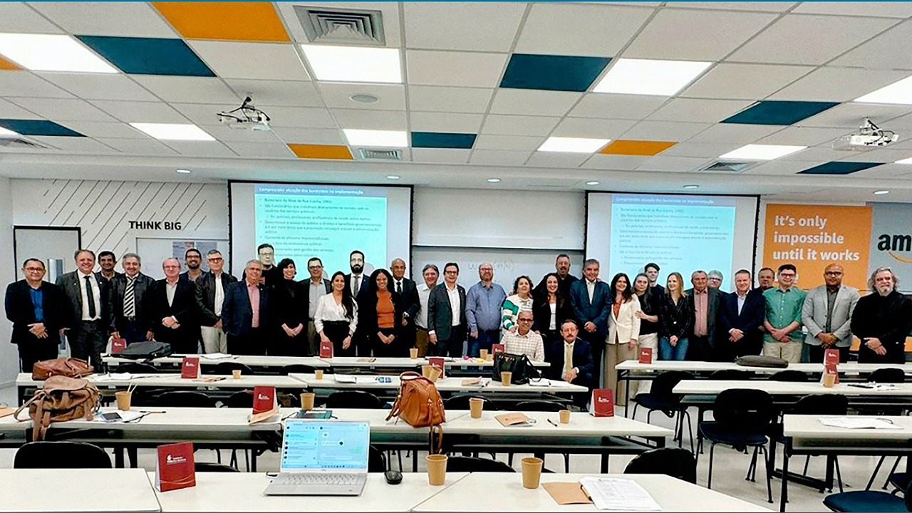 Foto de 35 participantes do curso — 8 mulheres e 27 homens, entre eles, o conselheiro Aderson Flores. A maioria das pessoas está em pé — apenas dois homens estão sentados. À frente deles, mesas e cadeiras e, ao fundo dois telões. 