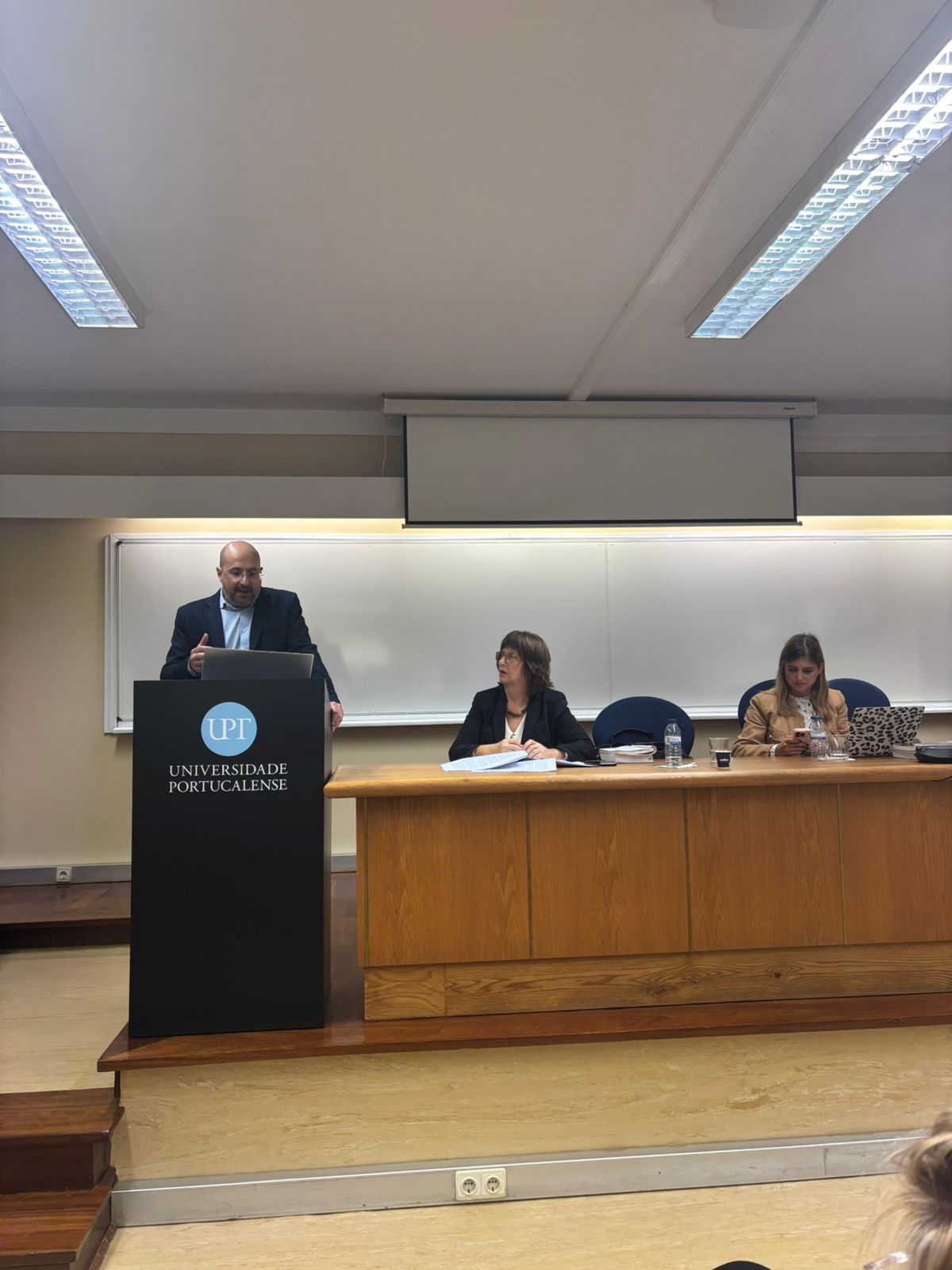 Foto do auditor fiscal de Controle Externo Renato Bossle Miguel discursando no púlpito, na Universidade Portucalense. Ele está em pé, à esquerda, ao lado de outras duas mulheres, que compõem uma bancada de autoridades. 