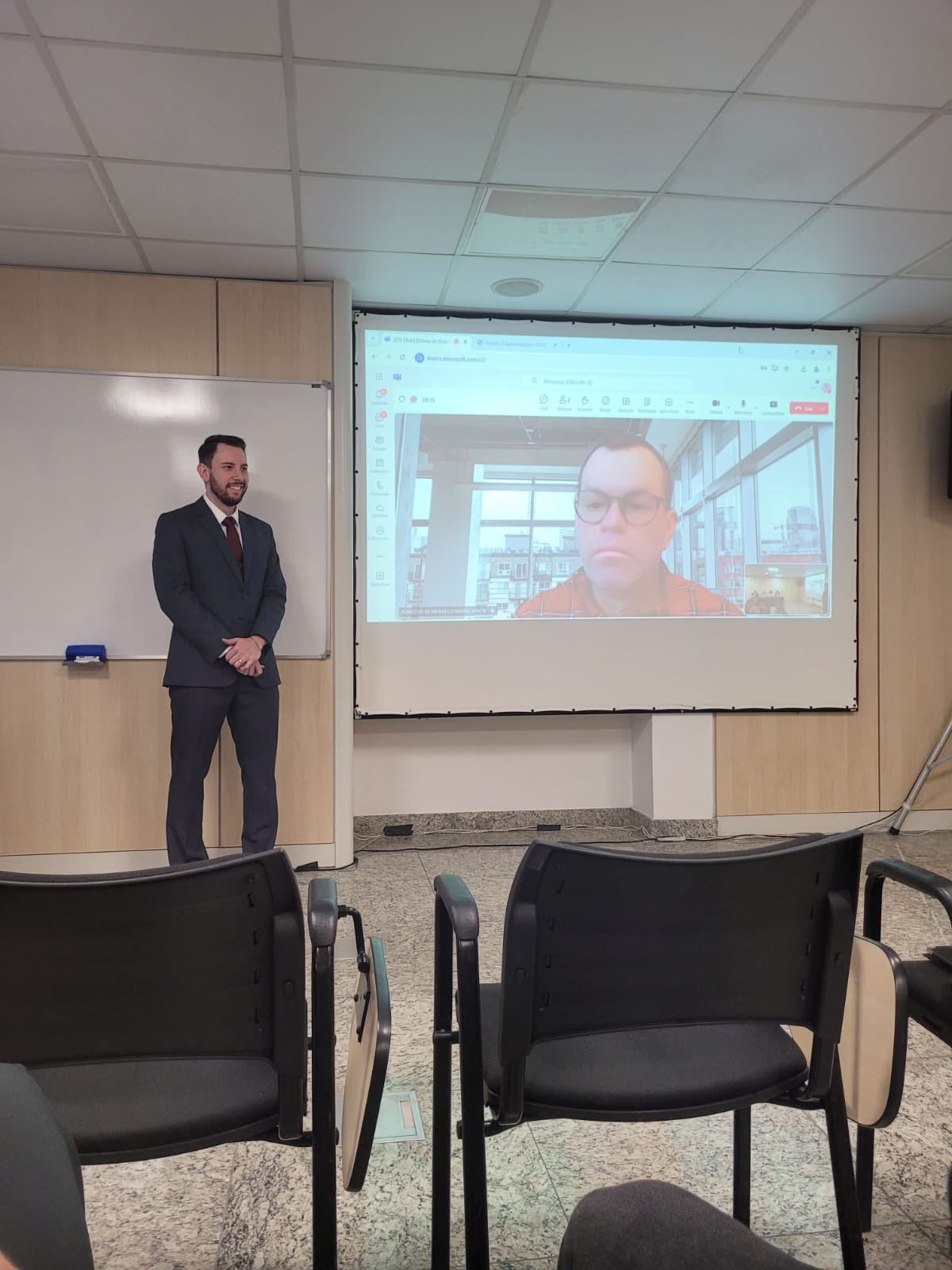 Foto ampla do auditor fiscal de Controle Externo Geovane Eziel Cardoso durante apresentação de mestrado. Ele está em pé, em uma sala do Icon. Ao lado, no telão, é exibida a imagem do conselheiro Adircélio de Moraes Ferreira Júnior, participando virtualmente.