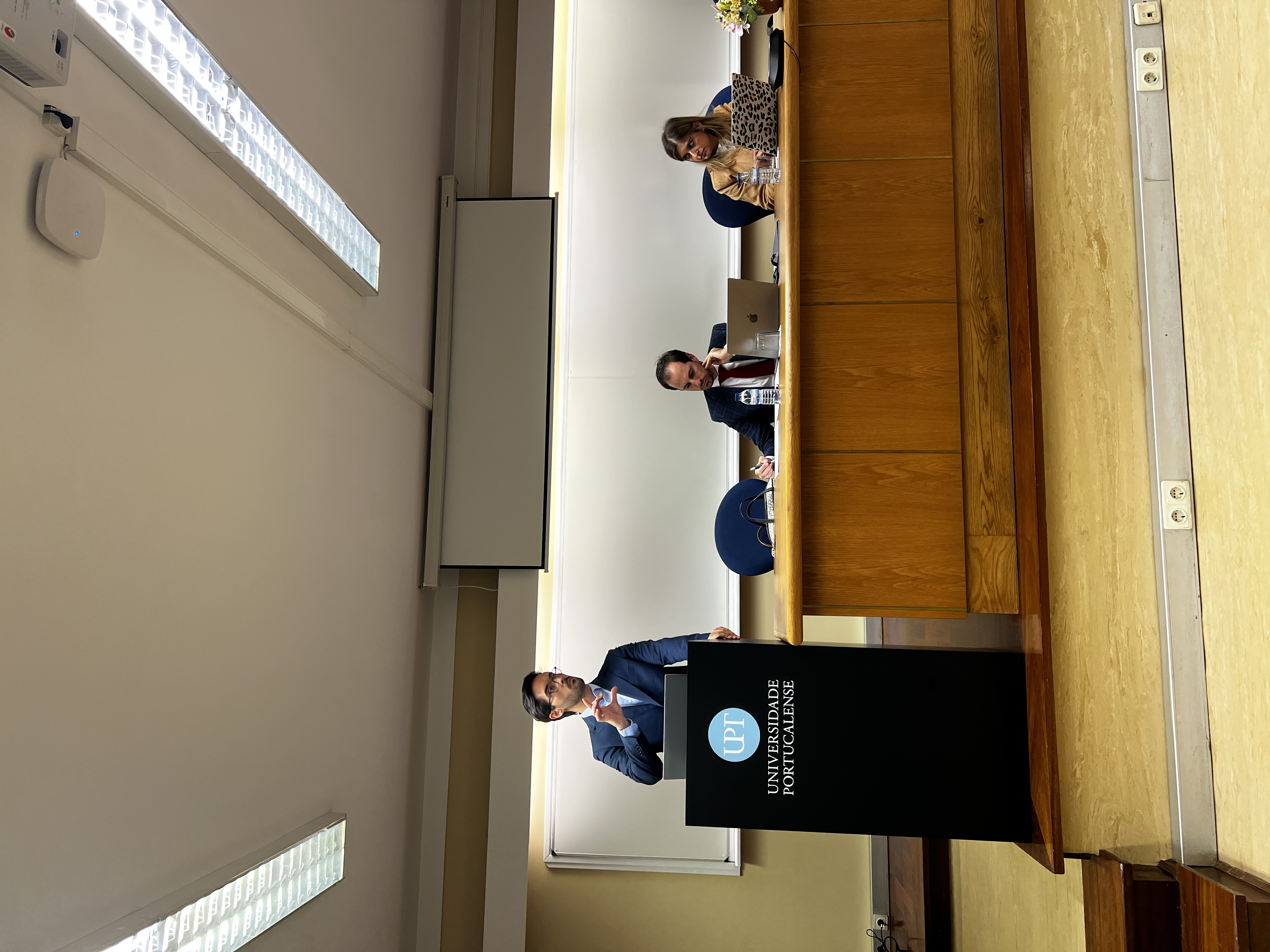 Foto do auditor fiscal de Controle Externo Juliano Frassetto Velho discursando no púlpito da Universidade Portucalense. Ele está em pé, ao lado da bancada de autoridades, diante da qual estão um homem e uma mulher. 