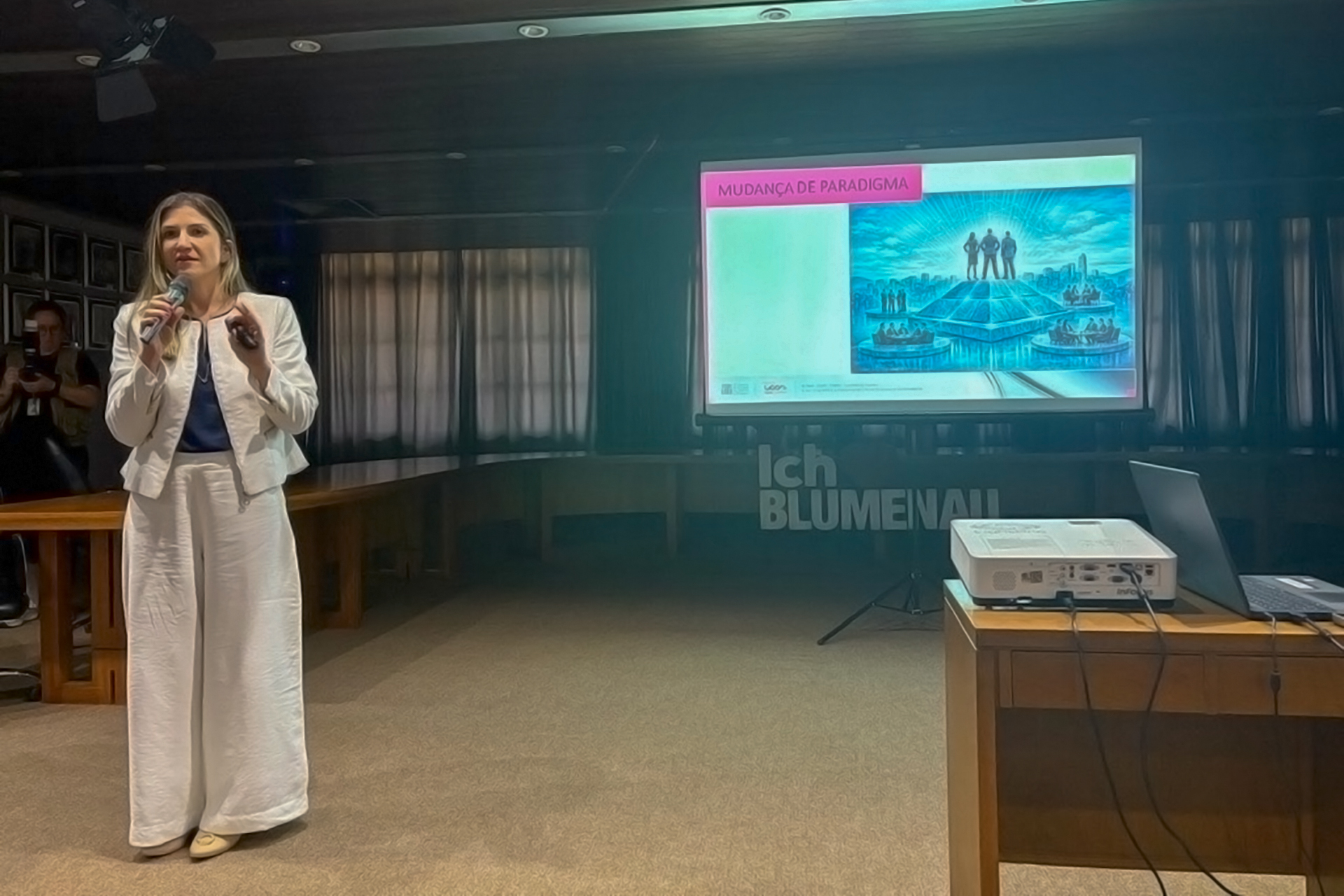Foto da servidora do TCE/SC Marina de Miranda falando ao público. Ela é uma mulher branca, de cabelos loiros. Veste roupa branca e está em pé. Ao fundo, telão projeta apresentação.