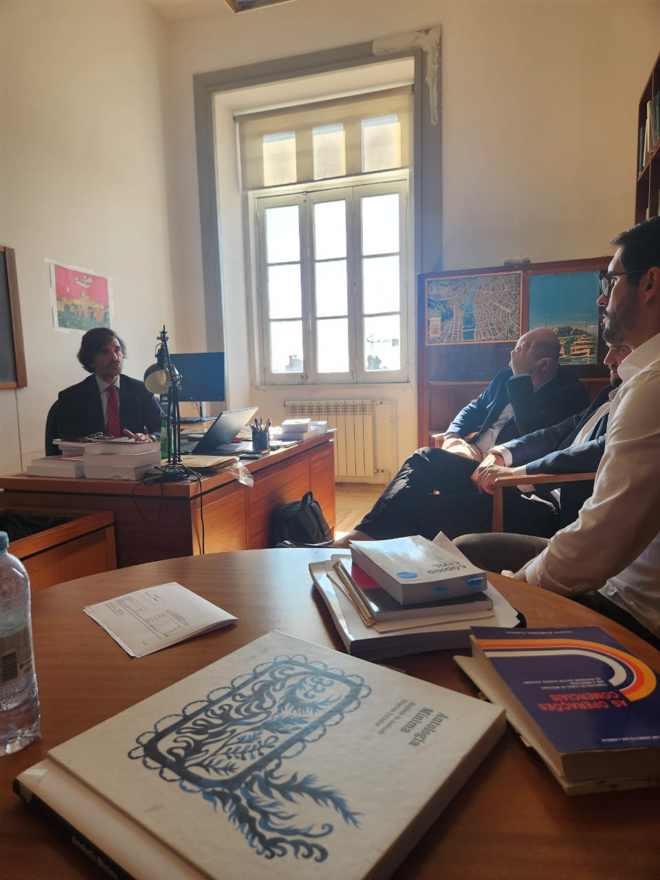 Foto com os auditores fiscais de Controle Externo Juliano Frasseto Velho, Renato Bossle Miguel e Rafael Scherb, na Universidade do Porto. Junto, está o juiz-conselheiro Luiz Miguel Pestana. A foto mostra também uma mesa com vários livros. 