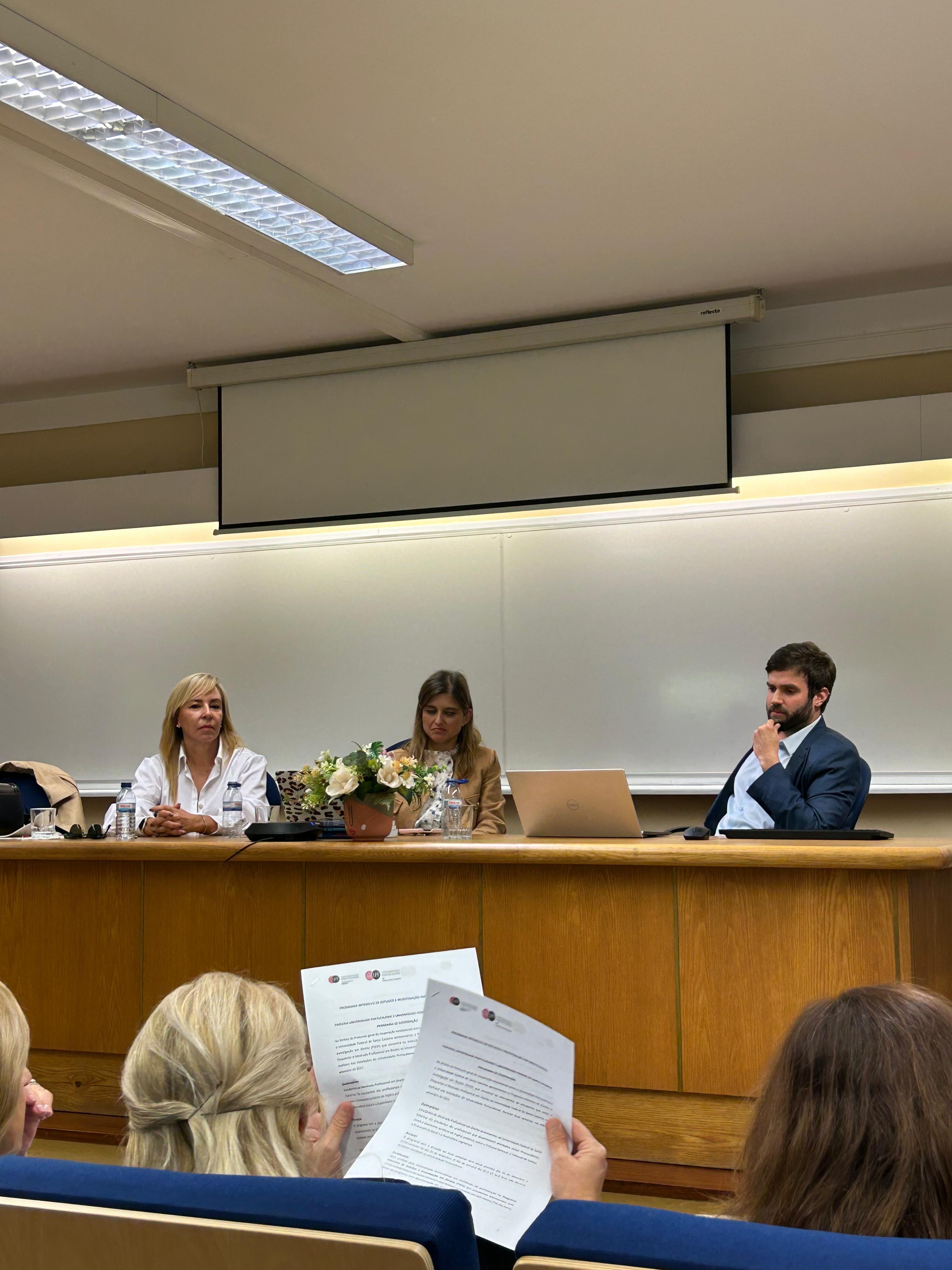 Foto do auditor fiscal de Controle Externo Rafael Scherb sentado diante de uma bancada, ao lado de outras duas mulheres, na Universidade Portucalense. 
