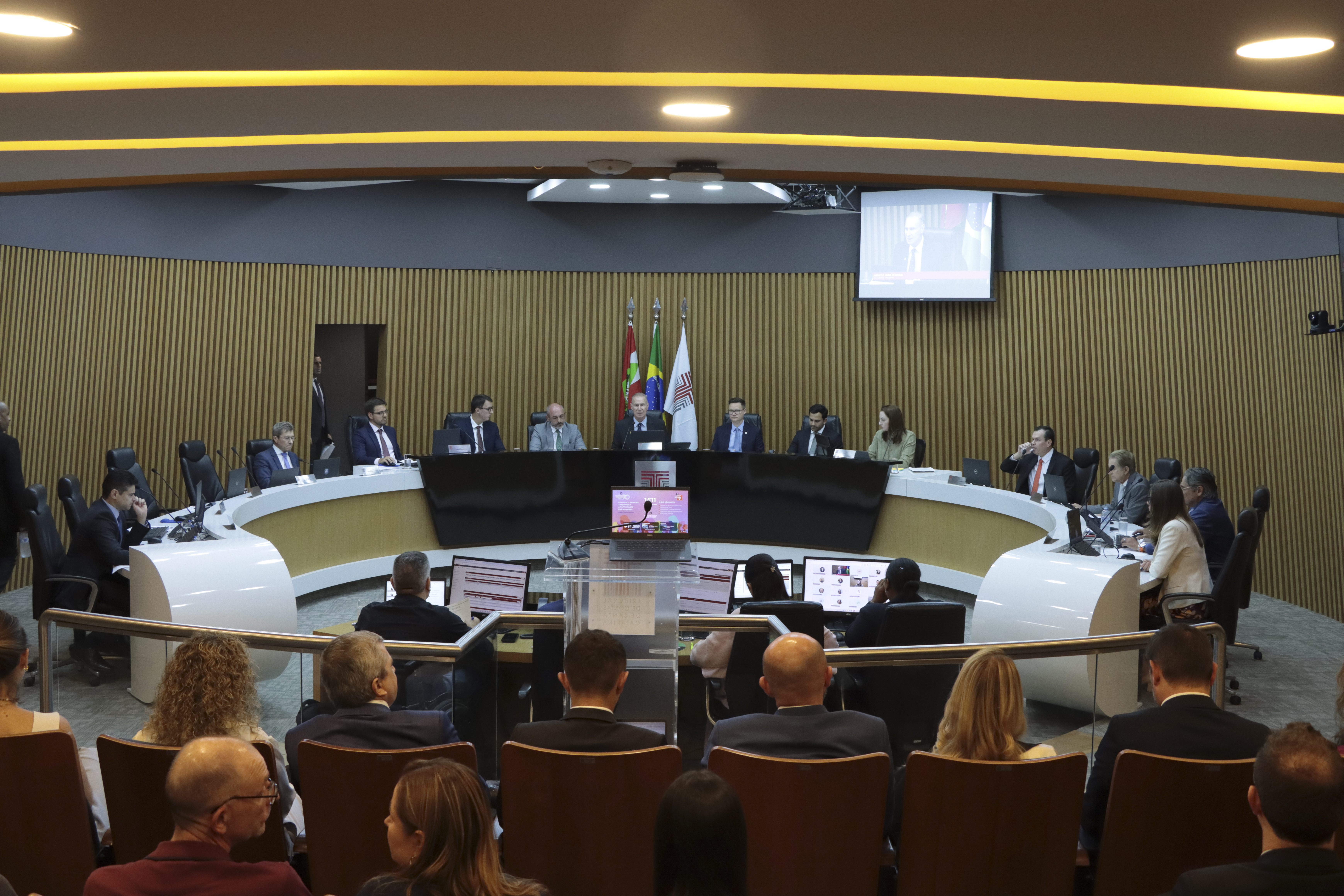 Foto horizontal do plenário do TCE/SC durante sessão, com autoridades sentadas em uma bancada semicircular ao centro, cada uma com computadores e documentos à frente. O ambiente é amplo, com painel de madeira ao fundo, bandeiras institucionais e iluminação no teto, enquanto ao primeiro plano aparecem participantes e público acompanhando a reunião.