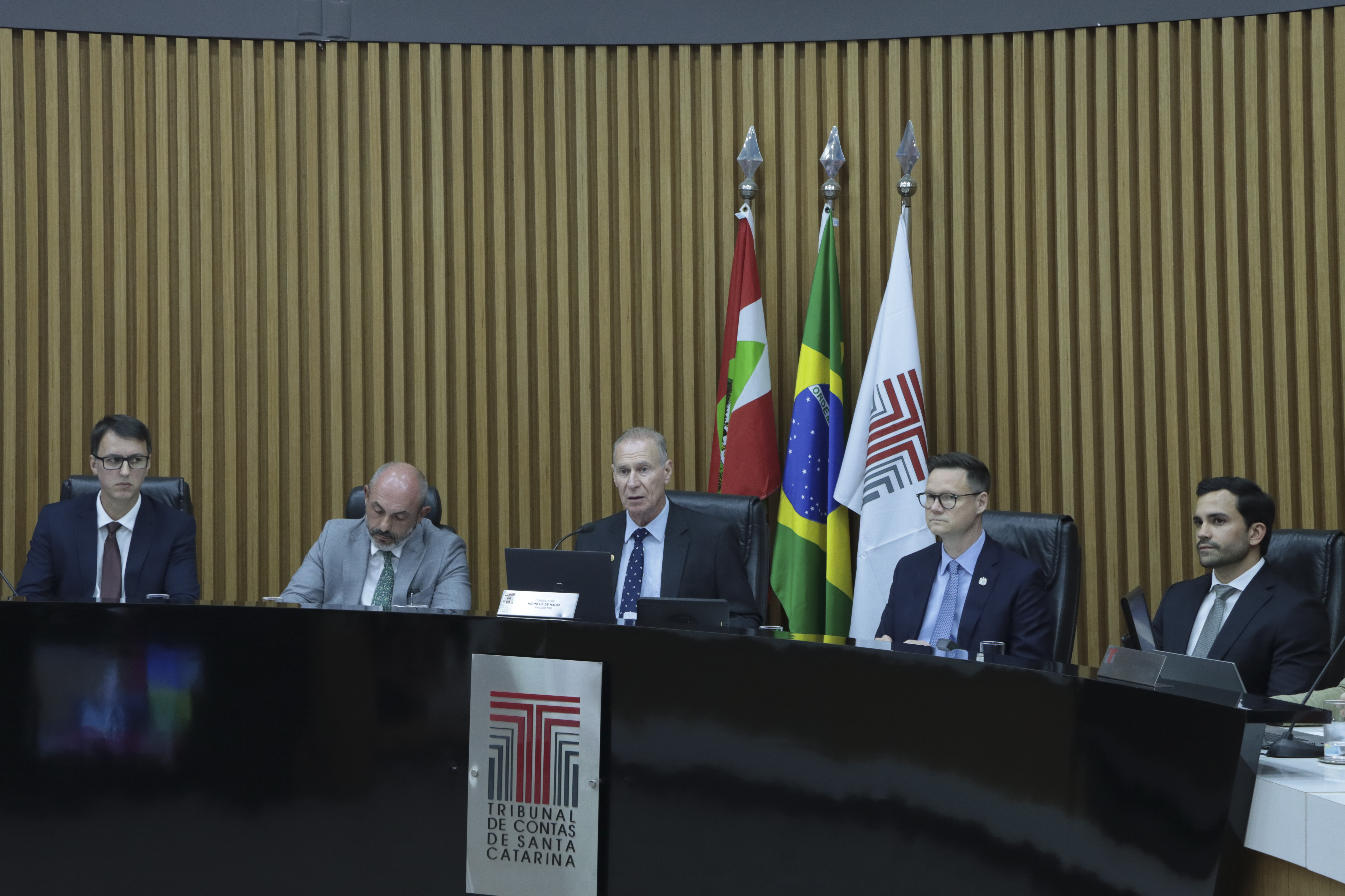 Foto horizontal da bancada do plenário do TCE/SC, com autoridades sentadas lado a lado em frente a uma mesa com computadores e documentos. Ao fundo, há painel de madeira e as bandeiras de Santa Catarina, do Brasil e do Tribunal. No centro da imagem estão o presidente do TCE/SC, conselheiro Herneus De Nadal, falando aos demais, o defensor público-geral Ronaldo Francisco, o secretário da Fazenda Cleverson Siewert e o desembargador André Luiz Dacol.