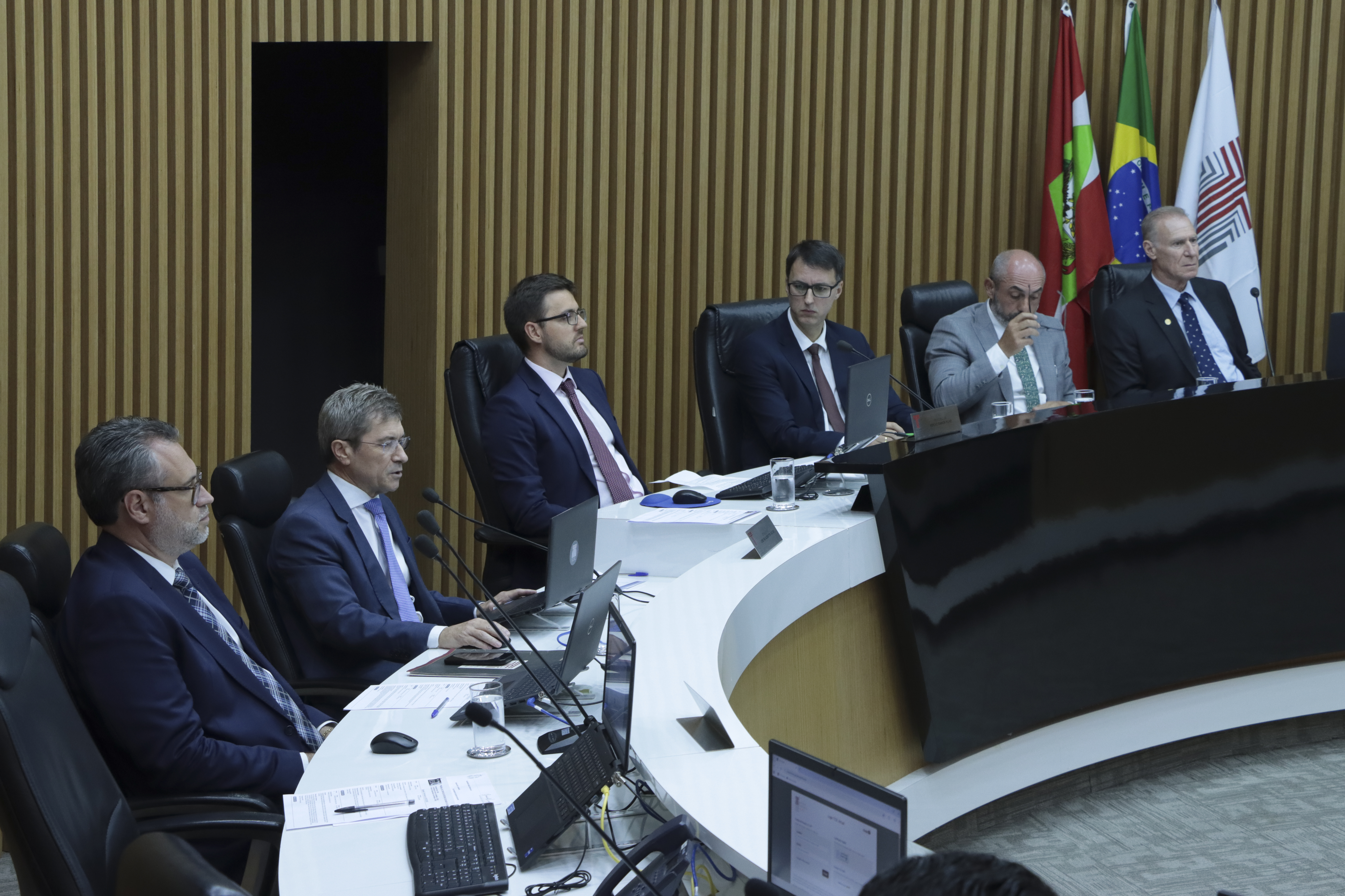 Foto horizontal do plenário do TCE/SC com autoridades sentadas em bancada curva, utilizando computadores e documentos durante a sessão. Em destaque, o vice-presidente José Nei Ascari, relator e propositor da mesa de consensualismo, fala ao público enquanto os demais acompanham atentos. Ao fundo, há painel de madeira e bandeiras institucionais, compondo o ambiente formal.