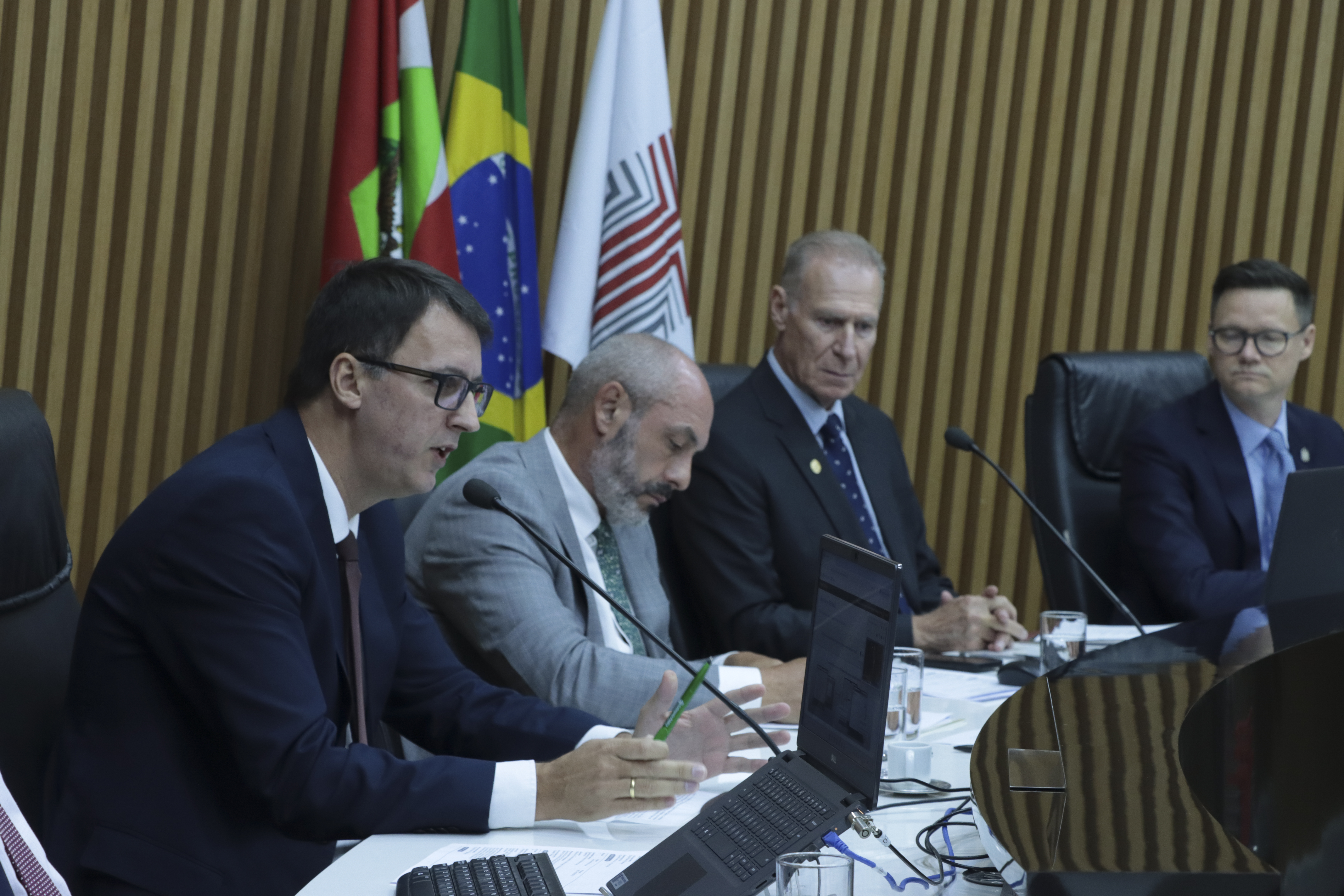 Foto horizontal da bancada do plenário do TCE/SC, com autoridades sentadas lado a lado, utilizando computadores e documentos durante a sessão. Em destaque, o defensor público-geral, Ronaldo Francisco, fala ao microfone, enquanto os demais acompanham atentos. Ao fundo, há painel de madeira e as bandeiras institucionais, compondo o ambiente formal.