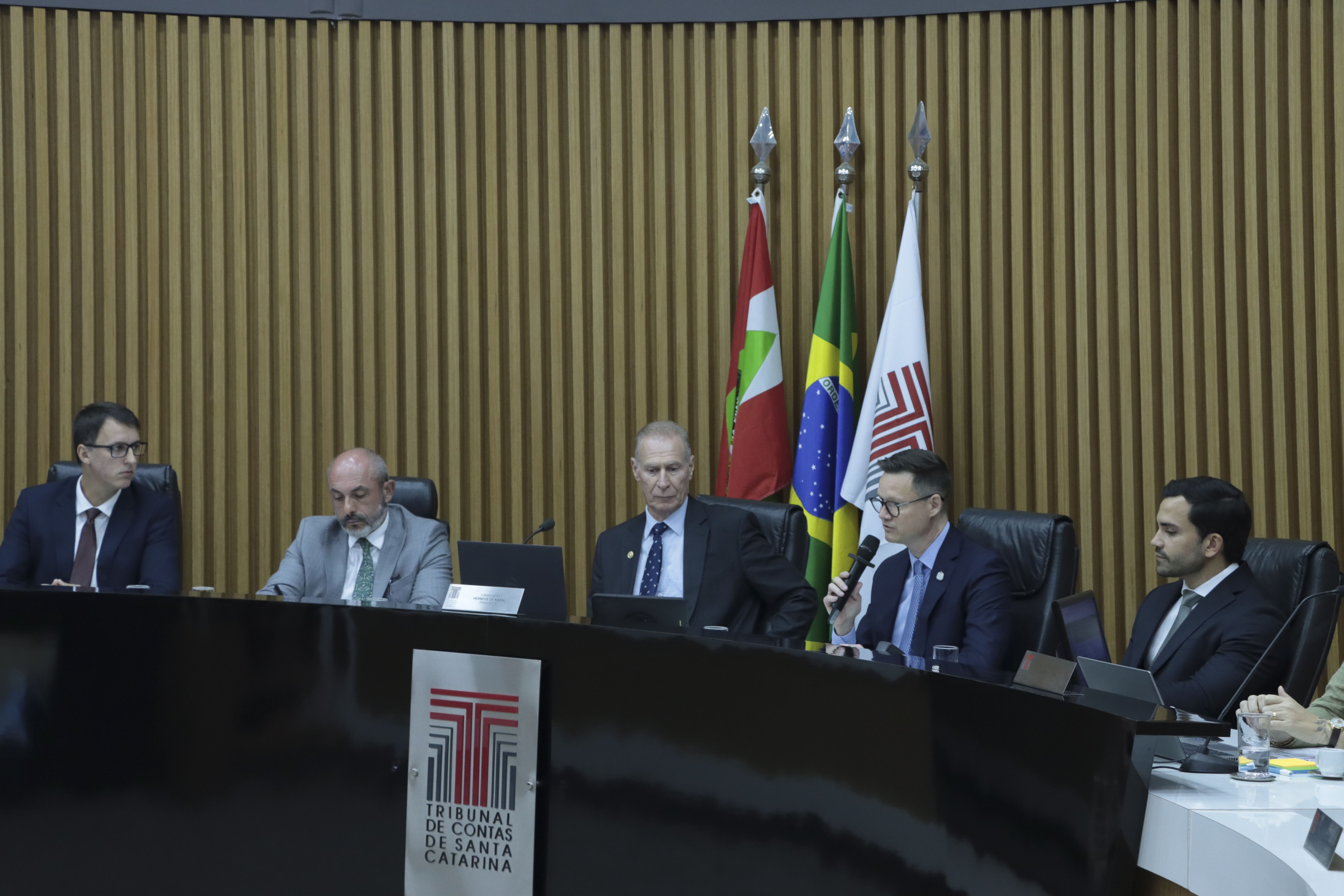 Foto horizontal da bancada do plenário do TCE/SC, com autoridades sentadas lado a lado, utilizando computadores e documentos durante a sessão. Em destaque, o secretário da Fazenda, Cleverson Siewert, fala ao microfone, enquanto os demais acompanham atentos. Ao fundo, há painel de madeira e as bandeiras institucionais, compondo o ambiente formal.