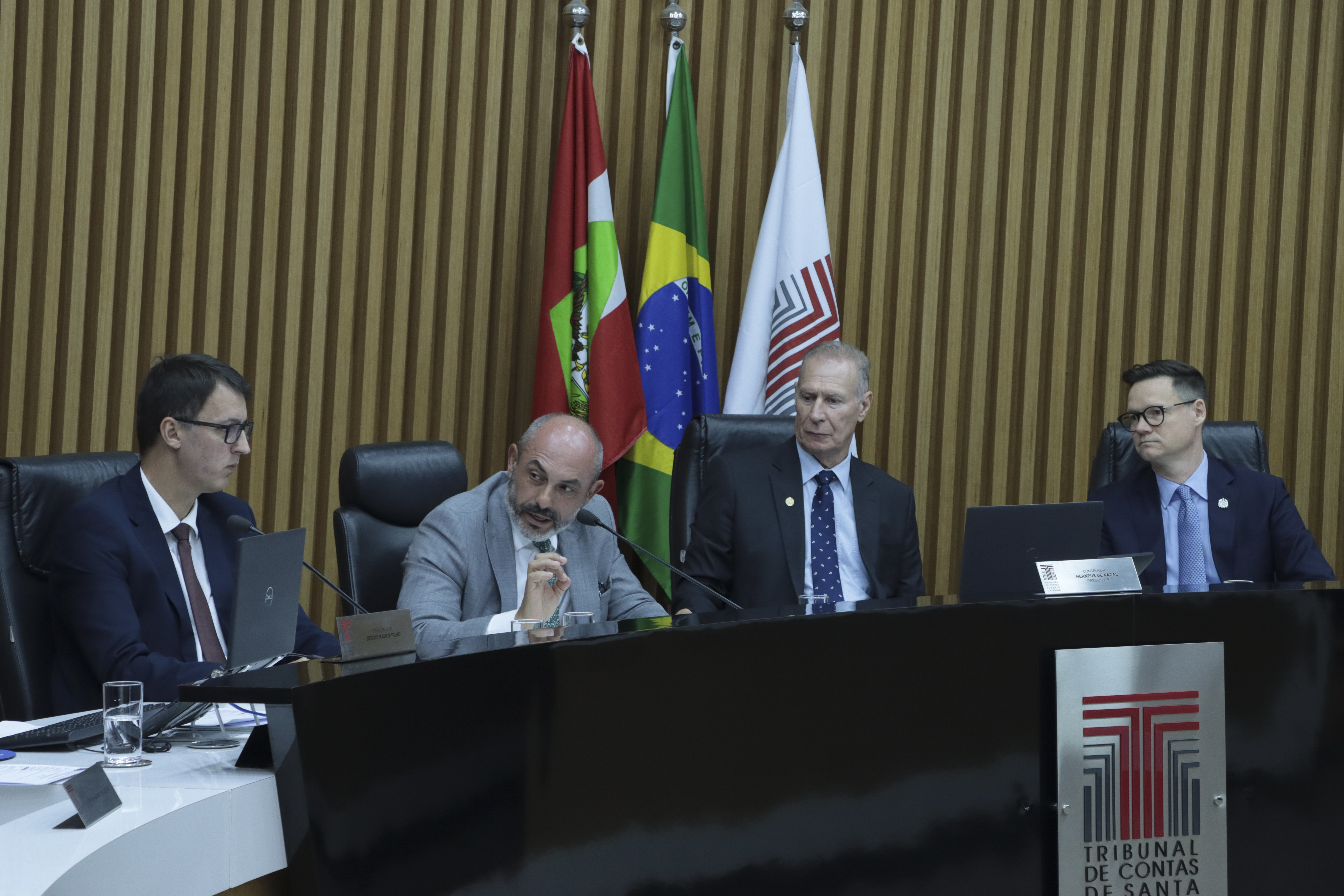 Foto horizontal da bancada do plenário do TCE/SC, com autoridades sentadas lado a lado, utilizando computadores e documentos durante a sessão. Em destaque, o desembargador André Luiz Dacol fala aos demais participantes, enquanto os outros acompanham atentos. Ao fundo, há painel de madeira e as bandeiras institucionais, compondo o ambiente formal.