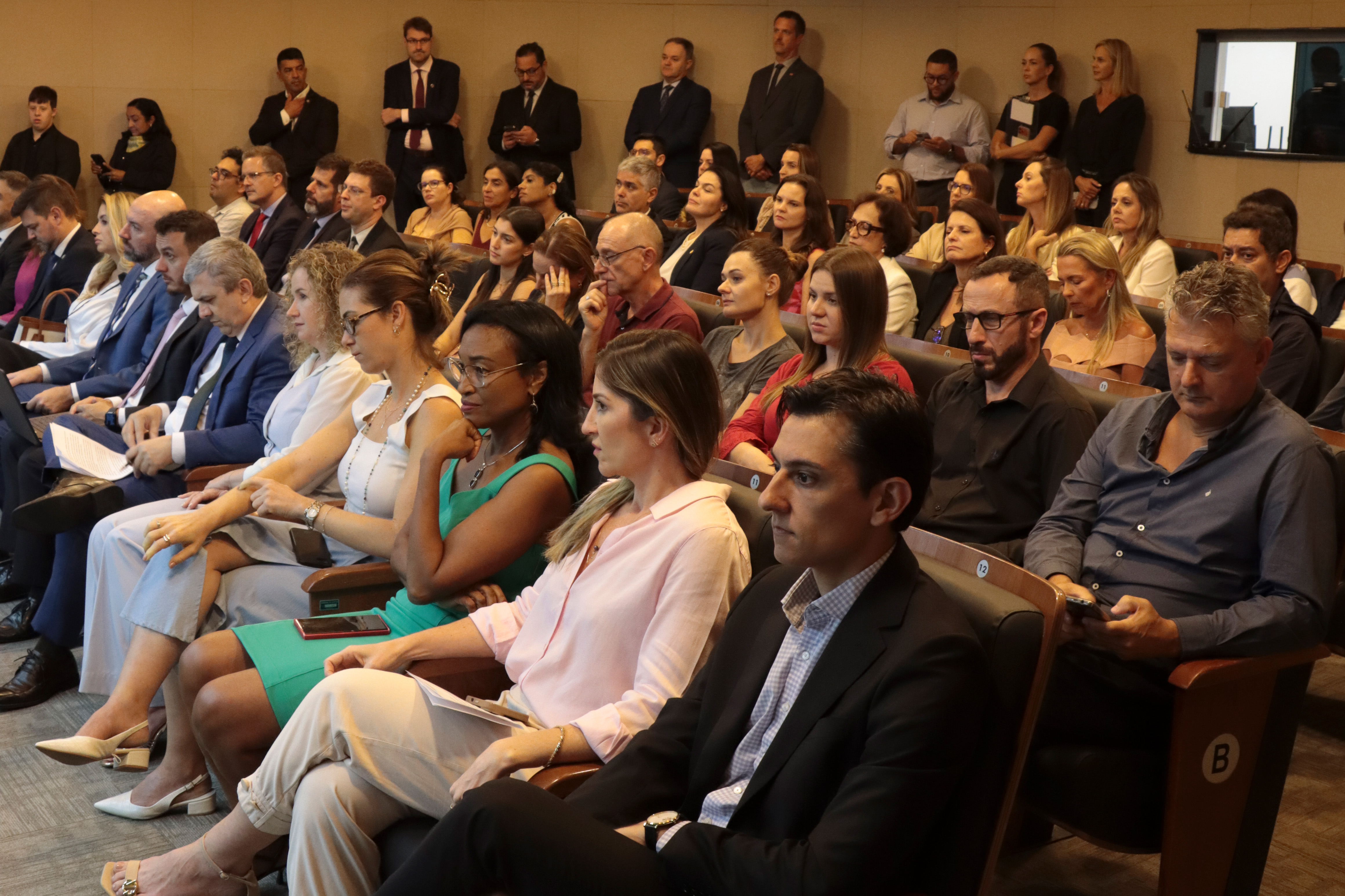Foto horizontal da plateia em auditório acompanhando a sessão no TCE/SC, com pessoas sentadas em fileiras de cadeiras estofadas, voltadas para a frente e atentas às falas. Entre os presentes, há servidores do Tribunal e outros participantes, alguns segurando documentos, cadernos ou celulares. Ao fundo, há mais pessoas em pé próximas à parede, também acompanhando a sessão, em um ambiente interno bem iluminado.