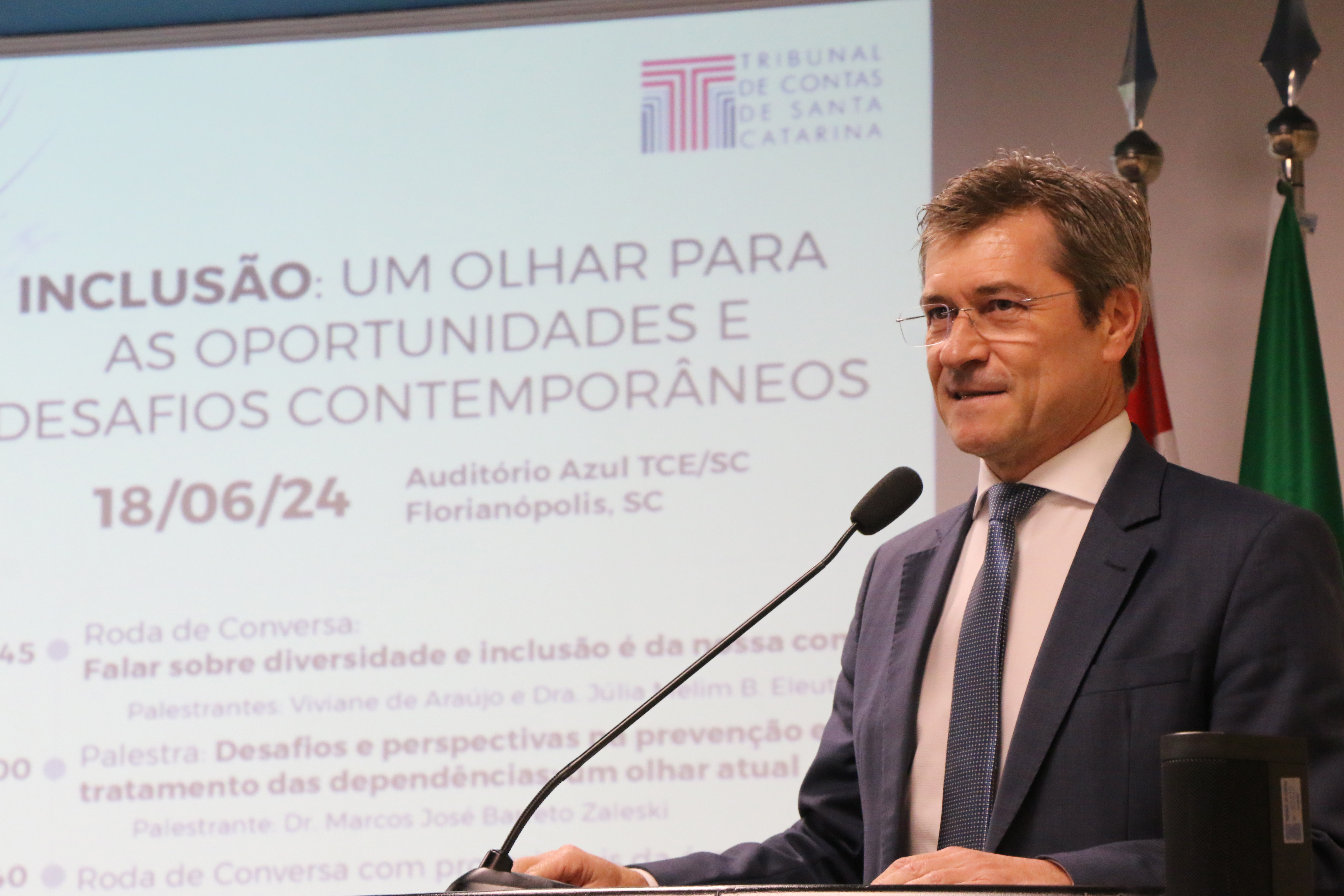 Banner horizontal com foto do conselheiro José Nei Ascari durante a abertura do evento. Ele é um homem branco, de cabelos claros. Veste terno e gravata e usa óculos de grau. Está em pé, diante do púlpito, falando ao microfone. Ao fundo, no telão, são projetadas as informações do evento. 