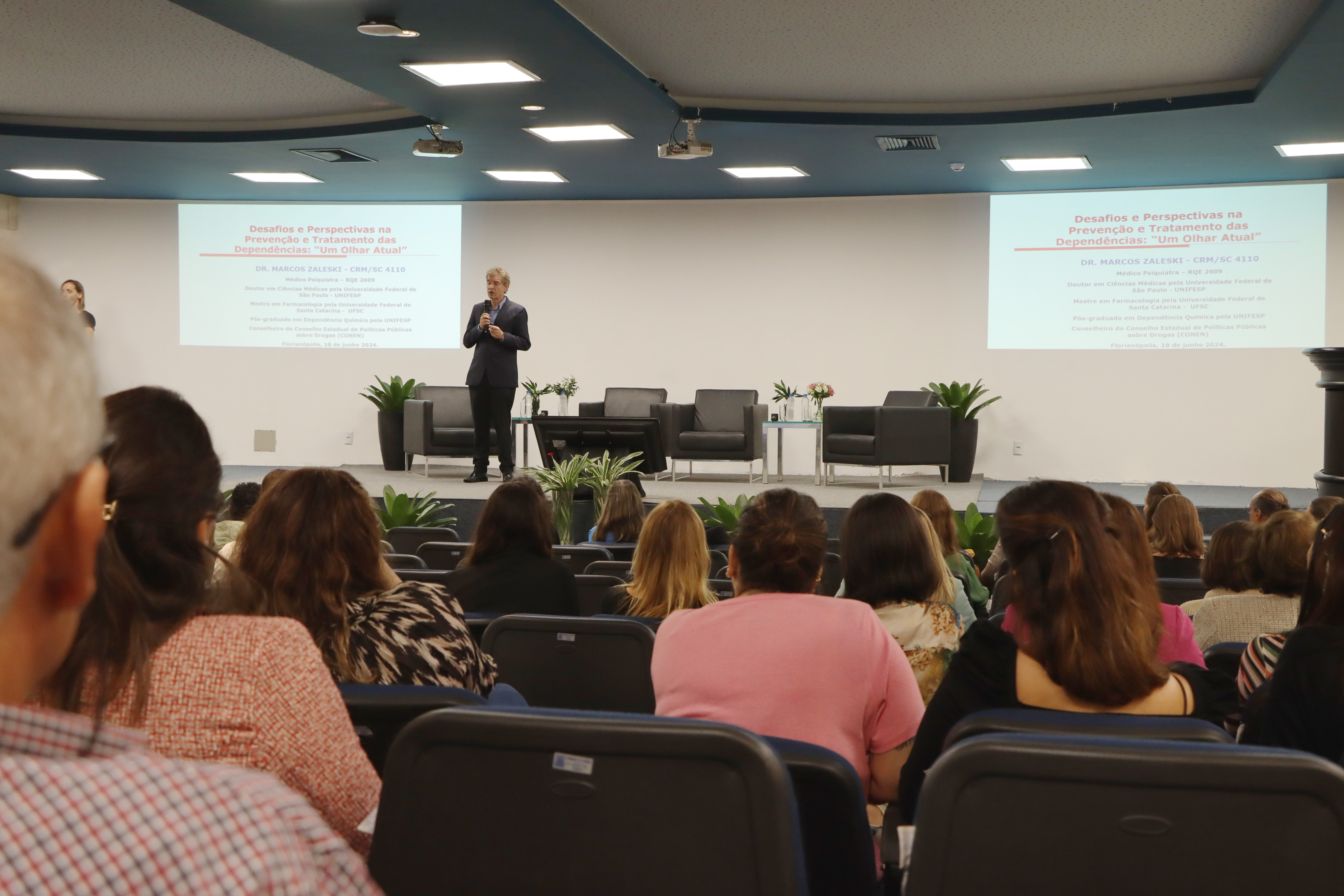 Foto dos palestrantes Marcos Zaleski, Cibele Lapolli, Nagele Queiroz e Valmor Júnior. Eles estão sentados em poltronas pretas, no palco do auditório do TCE/SC. Nagele segura o microfone com uma das mãos enquanto fala. À sua frente, de costas para a foto, parte do público que assiste à palestra, sentado em poltronas azuis do auditório. 
