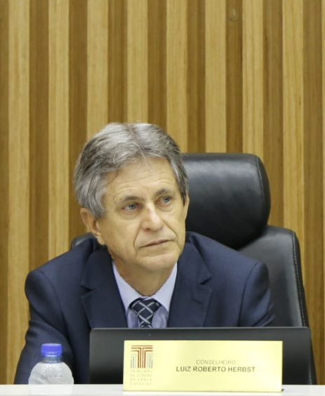 Fotografia do conselheiro Luiz Roberto Herbst, um homem de pele branca, cabelos grisalhos e olhos claros, olhando para o lado direito. Está sentado em uma cadeira preta, veste paletó azul-marinho e camisa azul-claro e usa gravata. À sua frente, um monitor de computador e uma placa na cor dourada com o seu nome.  