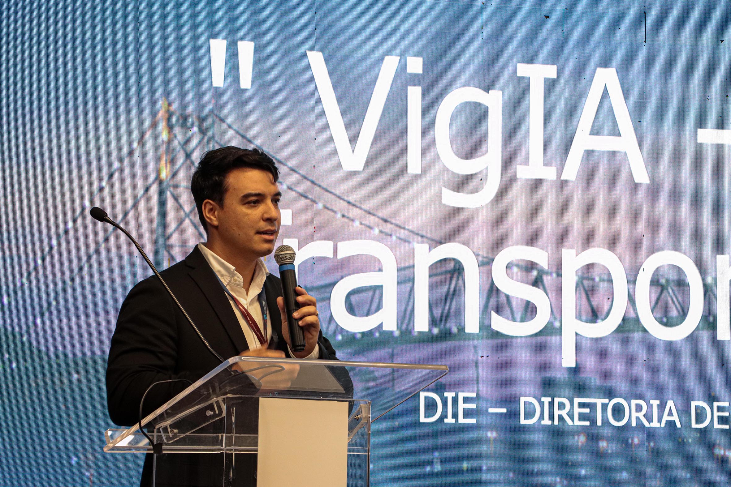 Foto do auditor fiscal de Controle Externo Lucas Favaretto falando ao microfone durante o CITC. Ele é um homem branco, de cabelos escuros. Veste paletó preto e está em pé, no palco, em frente ao telão que exibe a sua apresentação.