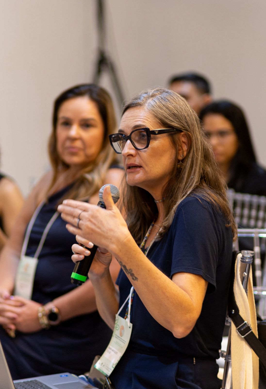 Foto da servidora Martha Godinho falando ao microfone durante sua participação no evento. Ela é uma mulher branca, de cabelos grisalhos. Veste roupa azul-escuro e usa óculos de grau. Está em meio aos demais participantes.
