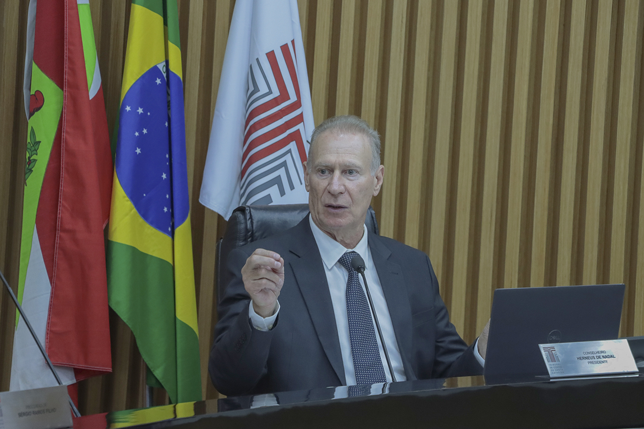 Foto horizontal do presidente do TCE/SC, conselheiro Herneus De Nadal, durante sessão plenária. Ele é um homem branco, de cabelos claros. Veste terno e gravata e fala ao microfone, sentado diante da bancada de autoridades. Ao fundo, há bandeiras e um painel de madeira.
