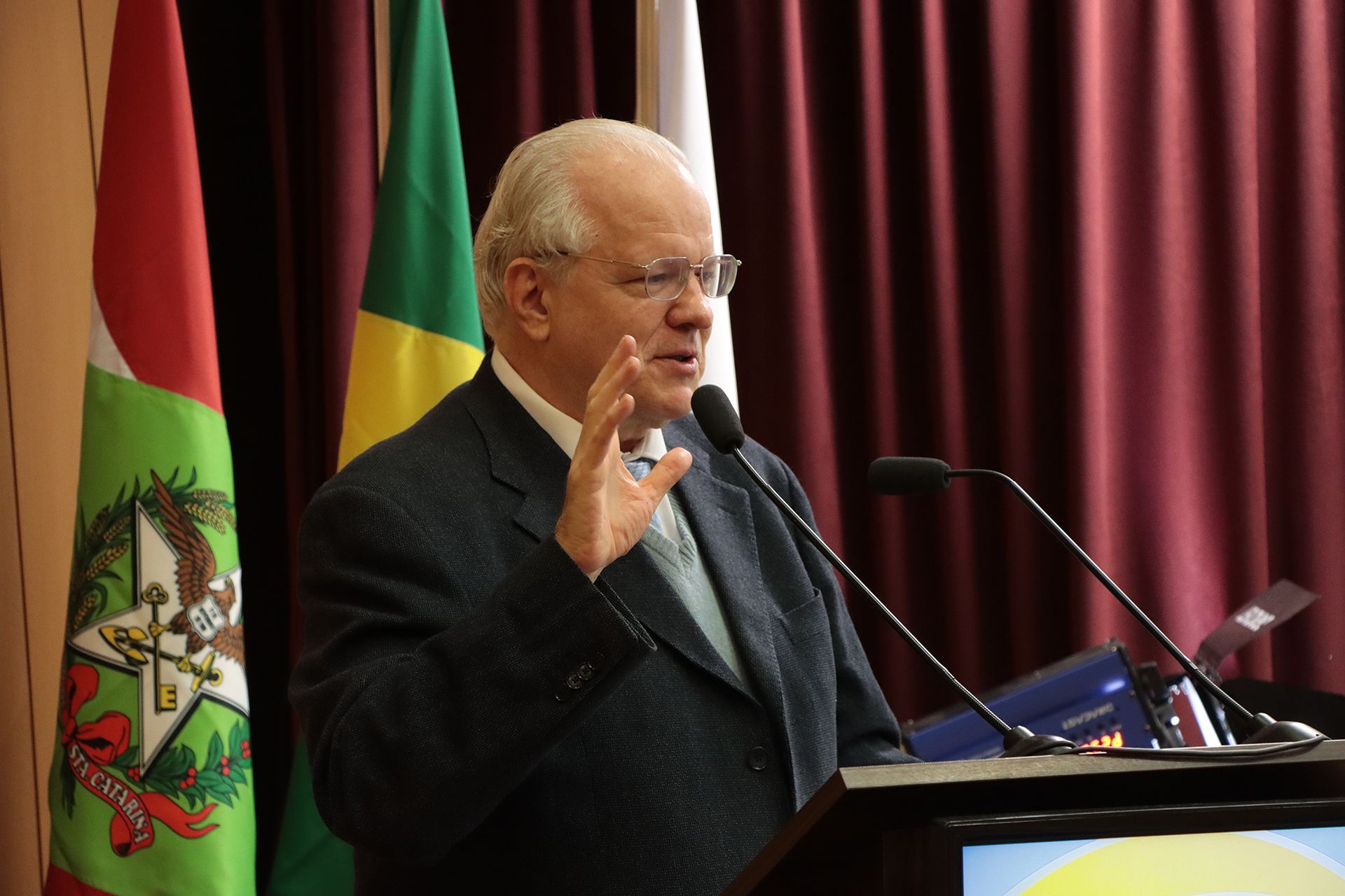 Foto do professor Juarez Freitas falando ao microfone no púlpito, durante painel. Ele é um homem branco, de cabelos grisalhos. Veste terno e gravata e usa óculos de grau. Gesticula enquanto fala. Ao fundo, cortinas bordôs.