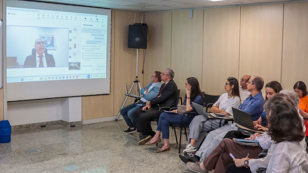 Foto dos participantes do debate sentados lado a lado, em sala no TCE/SC. No telão, fala uma das autoridades, de forma virtual. 