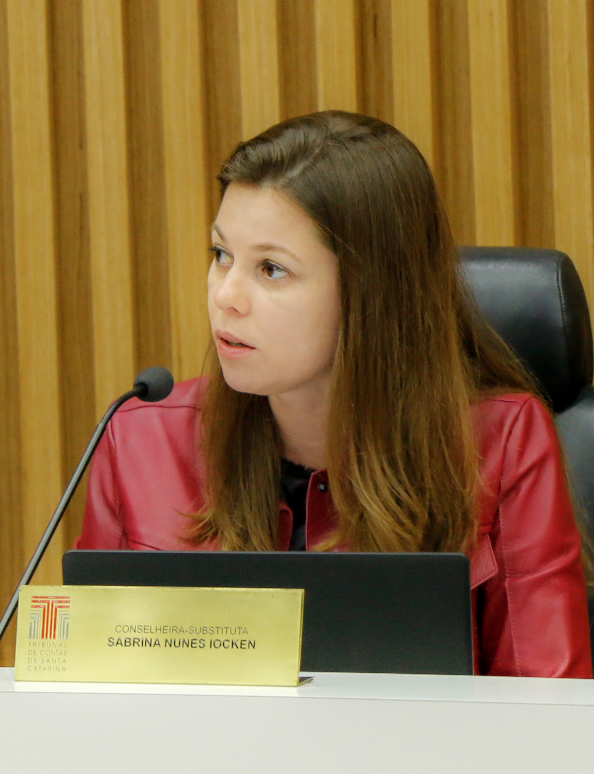 Fotografia da conselheira substituta Sabrina Nunes Iocken, uma mulher de pele branca e cabelos e olhos castanhos. Está olhando para a esquerda e sentada em uma cadeira preta. Veste casaco vermelho e blusa preta. À sua frente, um microfone, um monitor de computador e uma placa na cor dourada com o seu nome.  