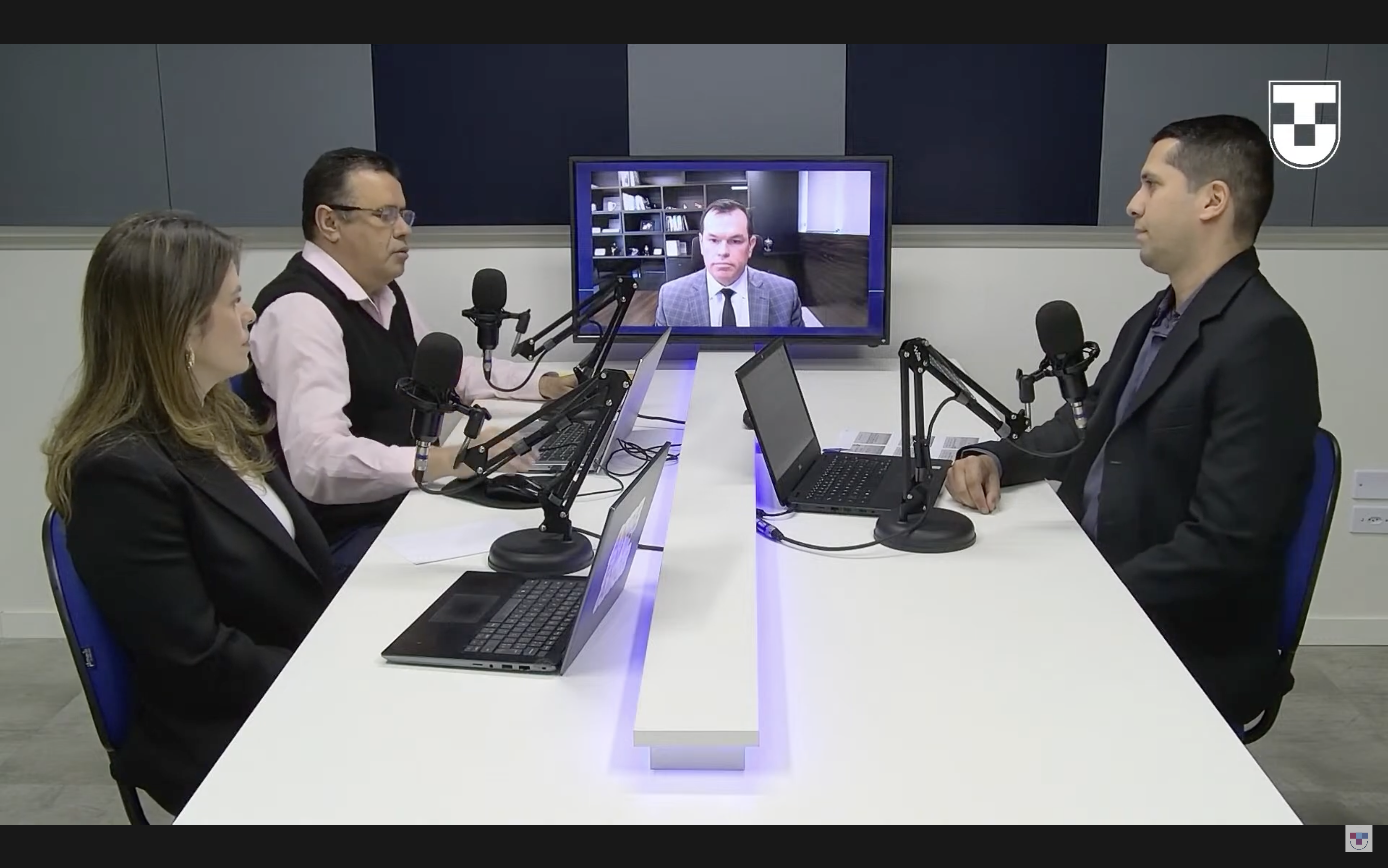 Banner horizontal com foto do pesquisador Rafael da Costa apresentando sua dissertação de mestrado. Ele está acompanhado dos professores Edson Vieira e Andréia Maricato. Ambos estão sentados diante de uma mesa de reuniões. Na televisão, ao fundo, participa, de forma remota, o conselheiro Adircélio de Moraes Ferreira Júnior. No canto superior direito, logo da Unitau. 