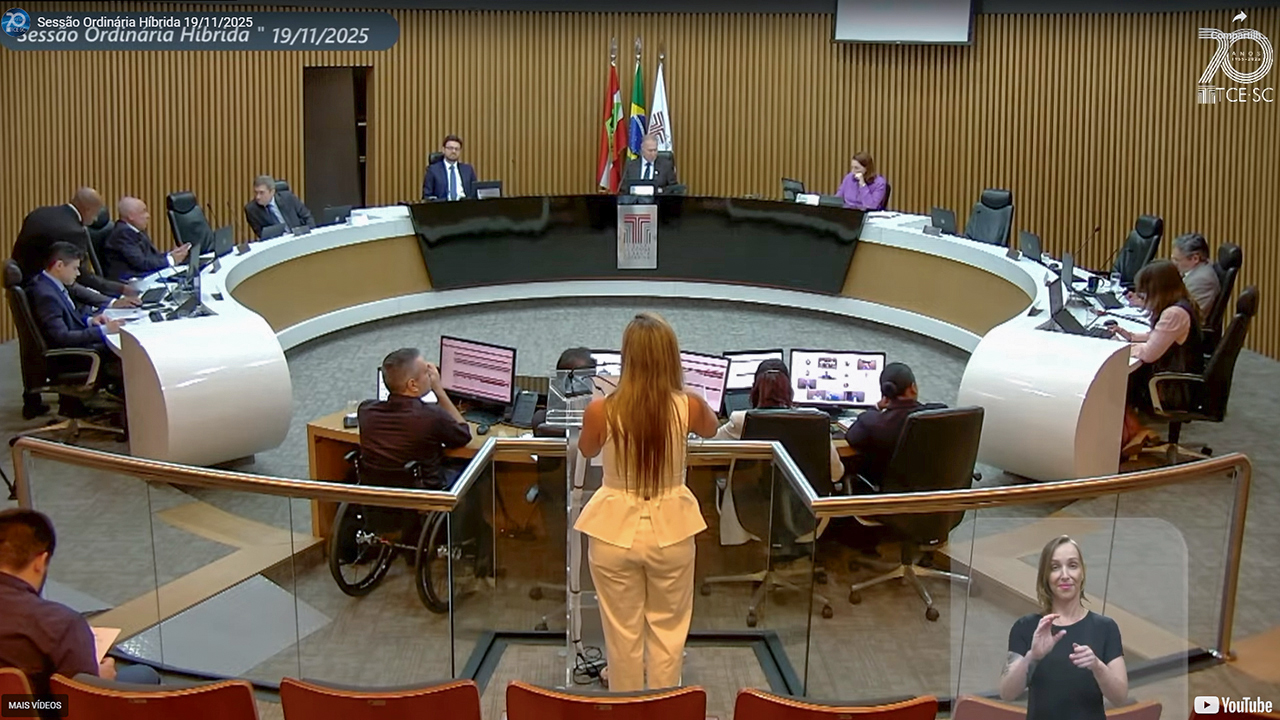 Foto ampla que mostra a bancada de autoridades do Plenário. Ao centro, o presidente, conselheiro Herneus De Nadal. À frente dele, em pé e de costas para a foto, falando ao público, a servidora Lúcia Helena. 