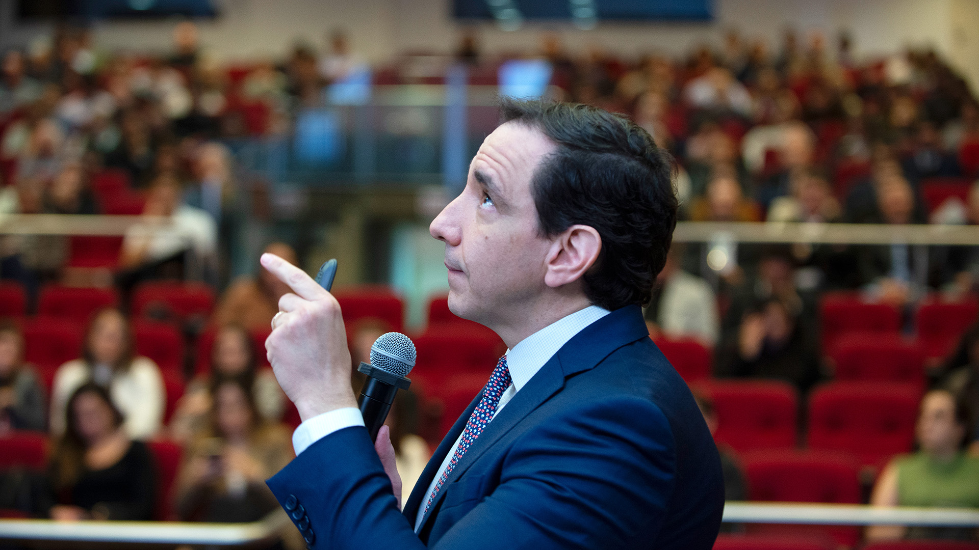 Foto horizontal e de perfil do diretor-presidente da ABCR, Marco Aurélio, durante palestra magna no Seminário Desenvolvimento e Infraestrutura. Ele é um homem branco, de cabelos pretos. Veste terno e gravata e aponta para cima. Ao fundo, desfocada, a plateia que o assiste, sentada em poltronas bordôs do auditório. 