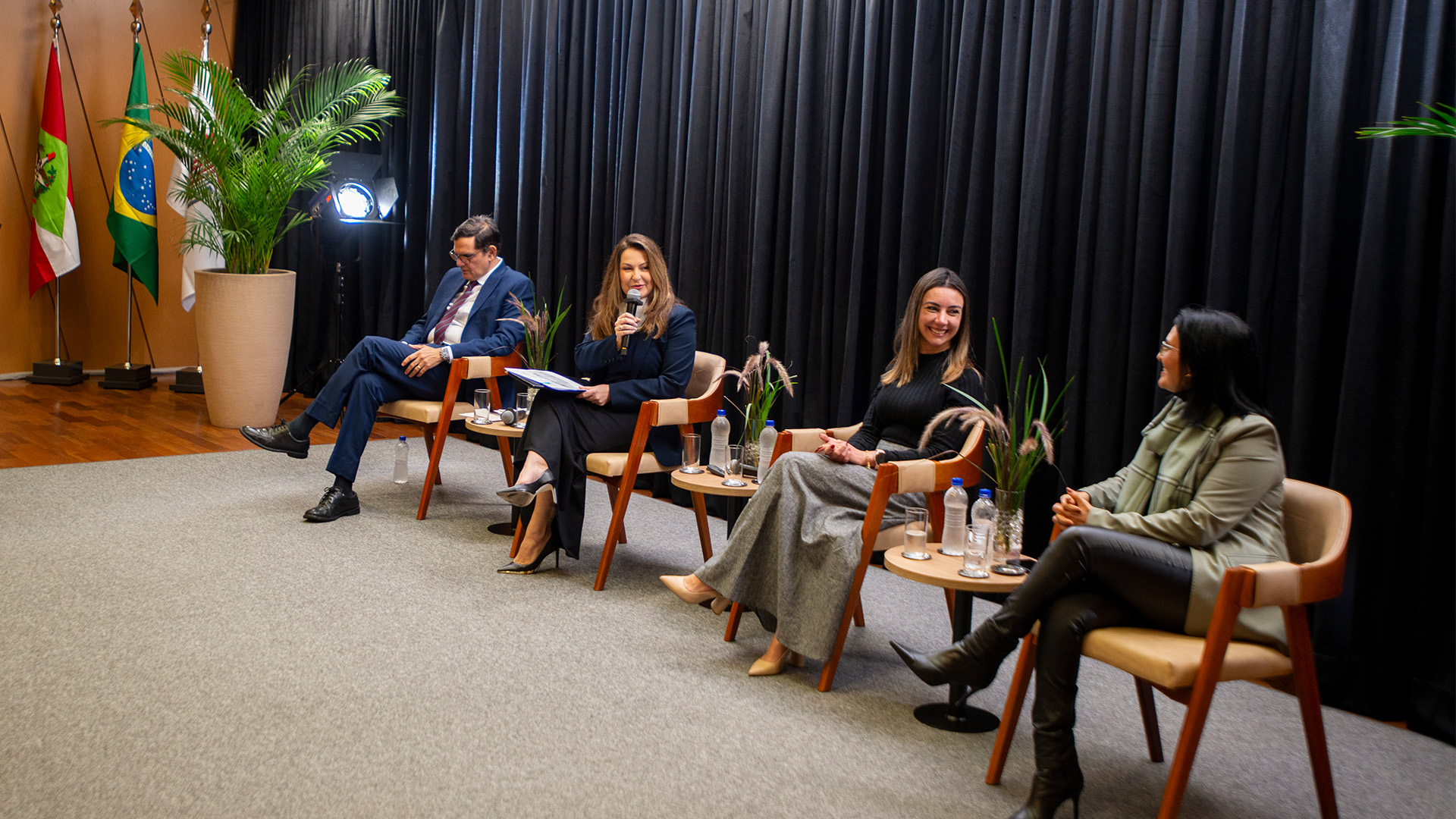 Foto horizontal dos integrantes da mesa sobre compras públicas. Entre os presentes, as auditoras fiscais de Controle Externo do TCE/SC Maira Galdino e Caroline de Souza, a gerente de Processos Licitatórios da B3 S.A., Mônica Lanna, e o conselheiro substituto do TCE/PE Marcos Nóbrega. Eles estão sentados, lado a lado, em poltronas, no palco do auditório, enquanto discutem o tema. 