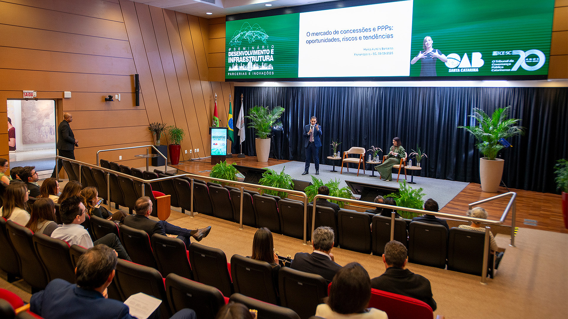 Banner horizontal com a imagem do diretor-presidente da Associação Brasileira de Concessionárias de Rodovias, Marco Aurélio de Barcelos Silva, durante palestra no Seminário Desenvolvimento e Infraestrutura. Ele é um homem branco, de cabelos escuros. Veste terno e gravata e está em pé, no palco do auditório do TCE/SC, enquanto fala ao público. Ao fundo, no telão, é projetada uma apresentação de slides e o logo do evento. 