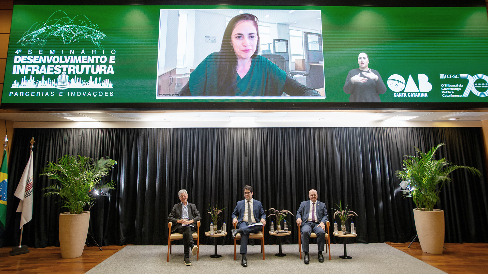 oto horizontal dos integrantes da mesa sobre mobilidade urbana. Entre os presentes, o presidente do Crea/SC, Carlos Xavier, o ex-superintendente da Suderf, Cassio Taniguchi, e o membro da Comissão da OAB/SC Rodrigo Lahoz. Eles estão sentados lado a lado, no palco do auditório do TCE/SC. Acima e ao fundo, no telão, é projetada a imagem da advogada Renata Dantas.