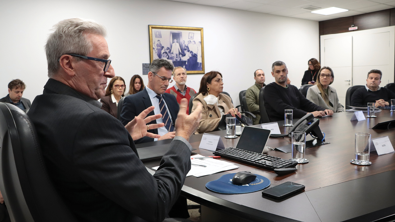 Foto horizontal de perfil do coordenador da mesa, Neimar Paludo, falando aos demais participantes. Há várias pessoas, representantes das demais instituições e servidores do TCE/SC, sentadas ao redor da mesa, ouvindo atentamente. 
