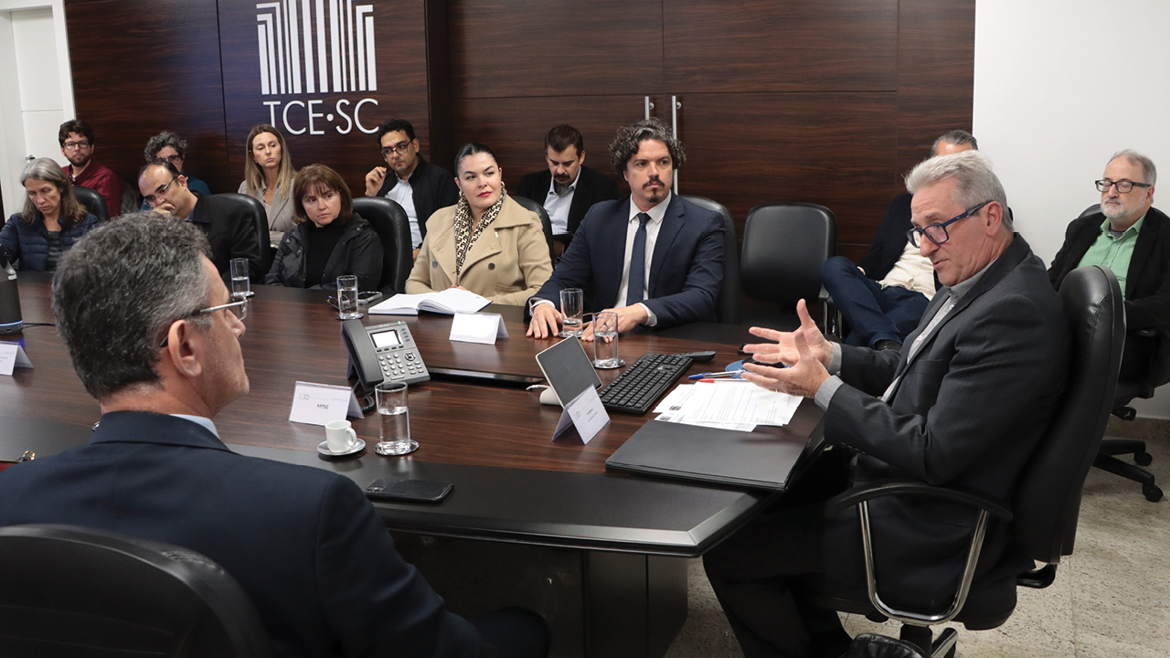 Foto horizontal que mostra outros participantes da mesa. Fala aos demais o coordenador, Neimar Paludo. Ao seu lado, o deputado estadual Marquito e a diretora-geral adjunta de Controle Externo, Monique Portella. Ao fundo, representantes de outras instituições. 