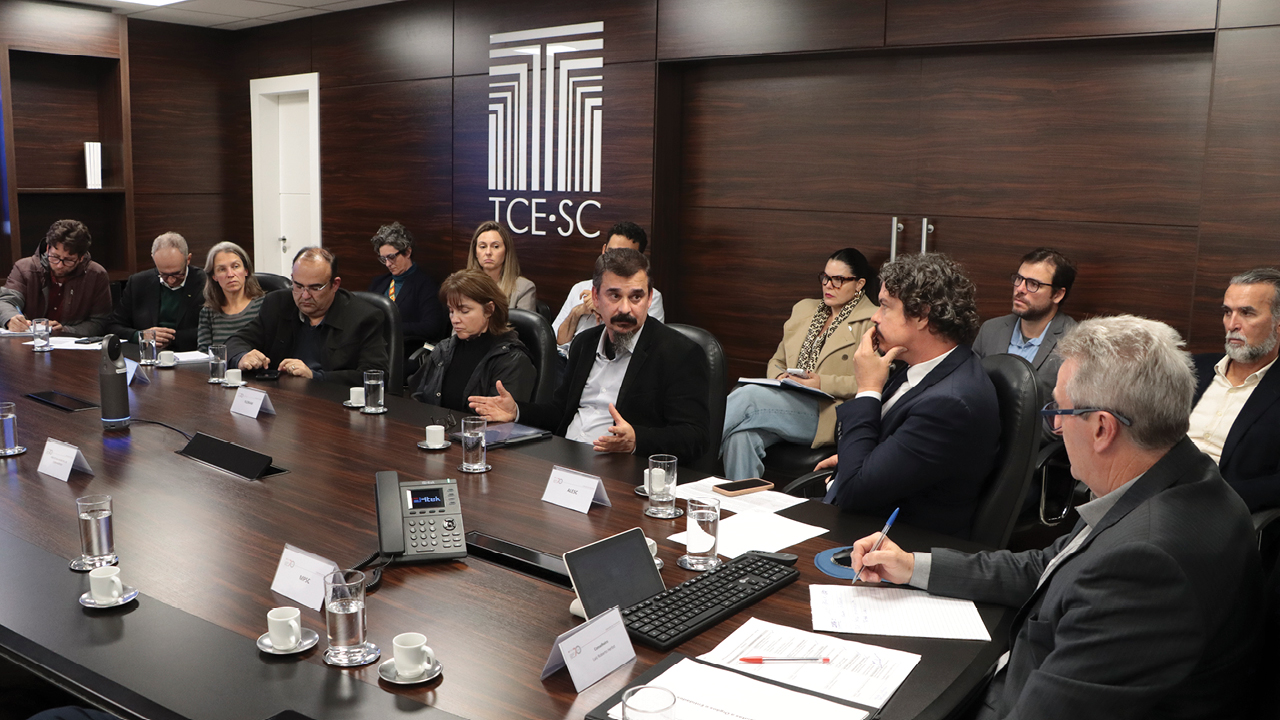 Foto que mostra vários participantes da mesa de consensualismo conversando. Entre os presentes, o coordenador, Neimar Paludo, o deputado Marquito e o professor da UFSC. Sobre a mesa diante da qual estão sentados, há papéis, canetas, copos de água e equipamentos eletrônicos. 