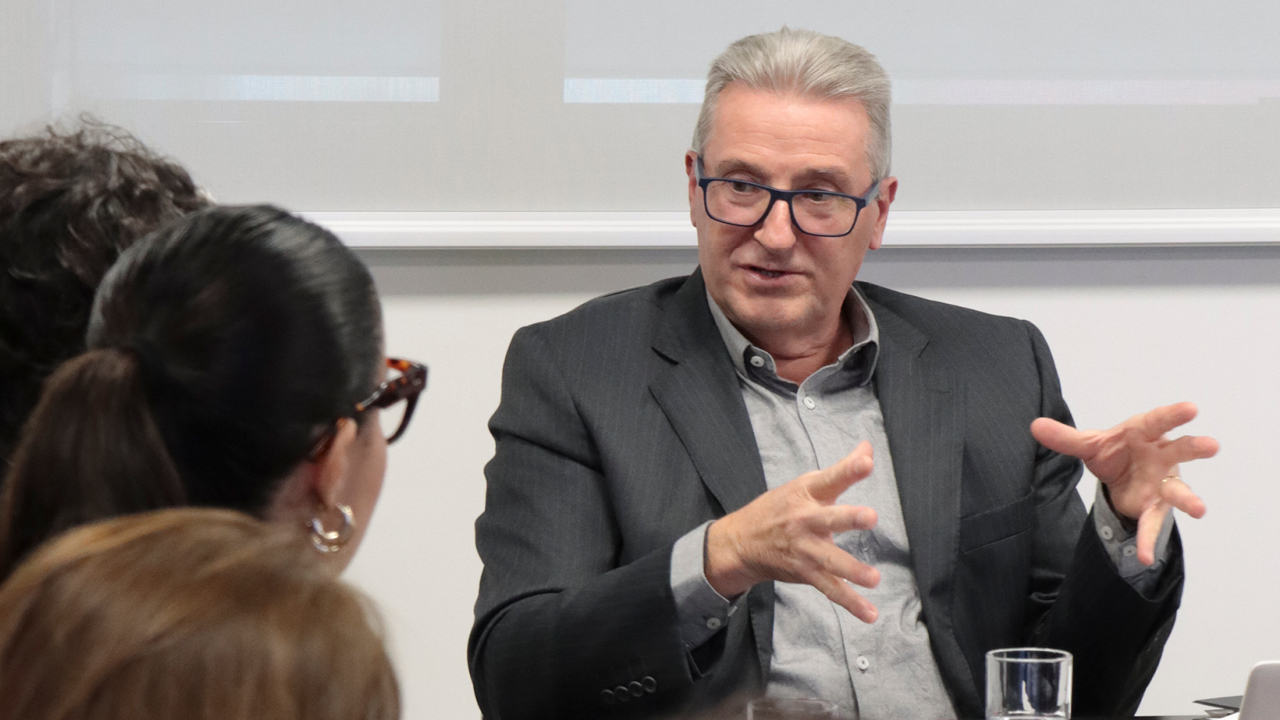 Foto do coordenador Neimar Paludo falando aos demais durante a reunião. É um homem branco, de cabelos grisalhos. Veste paletó preto e usa óculos de grau. Está gesticulando com as mãos. 