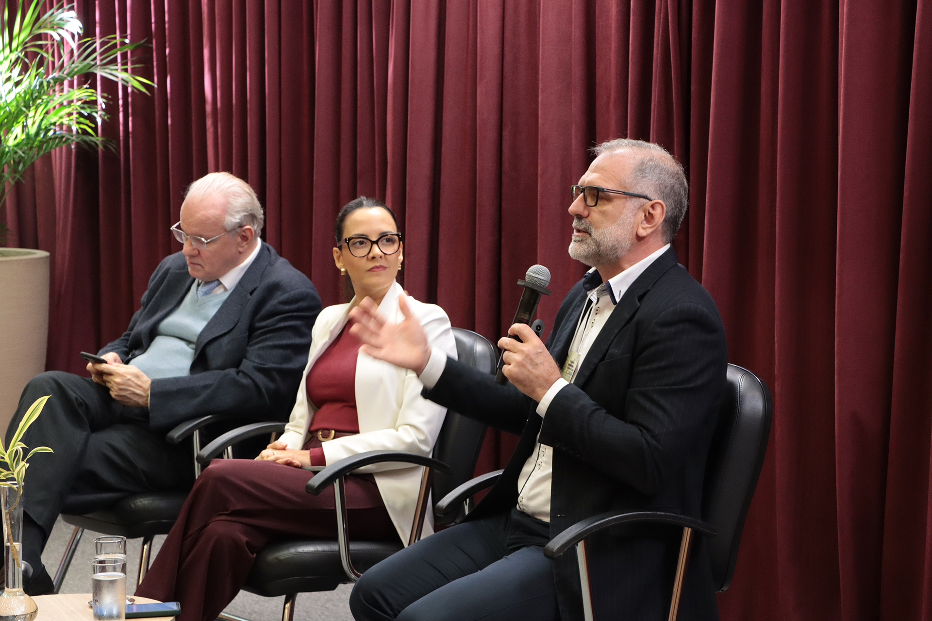 Foto do secretário de Agricultura Familiar e Agroecologia do MDA, Vanderley Ziger, falando ao microfone durante um dos painéis, no palco. É um homem branco, de cabelos e barba grisalhos. Veste paletó preto e está ao lado de outros dois palestrantes. Ao fundo, há cortinas bordôs.