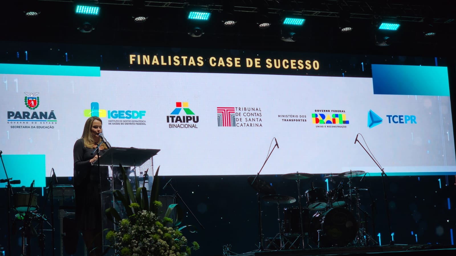 Foto com as logos dos finalistas do case de sucesso no telão: Governo do Estado do Paraná - Secretaria da Educação, IGESDF, Itaipu Binacional, TCE/SC, Ministério dos Transportes e TCE/PR. à frente, uma moça está falando do púlpito. 