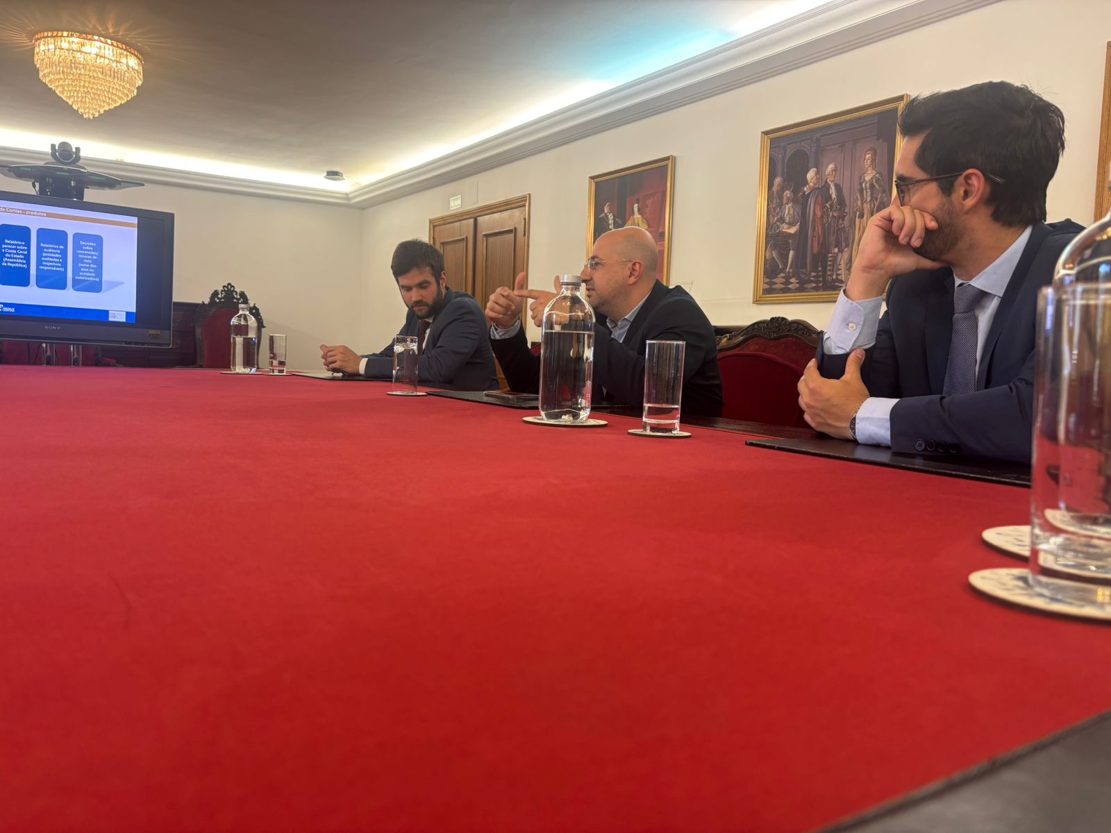Foto dos auditores fiscais de Controle Externo Rafael Scherb, Renato Bossle Miguel e Juliano Frassetto participando de reunião no Tribunal de Contas de Portugal. Eles estão sentados diante de uma mesa vermelha, enquanto conversam com os demais. Ao fundo, na parede, há quadros artísticos. 