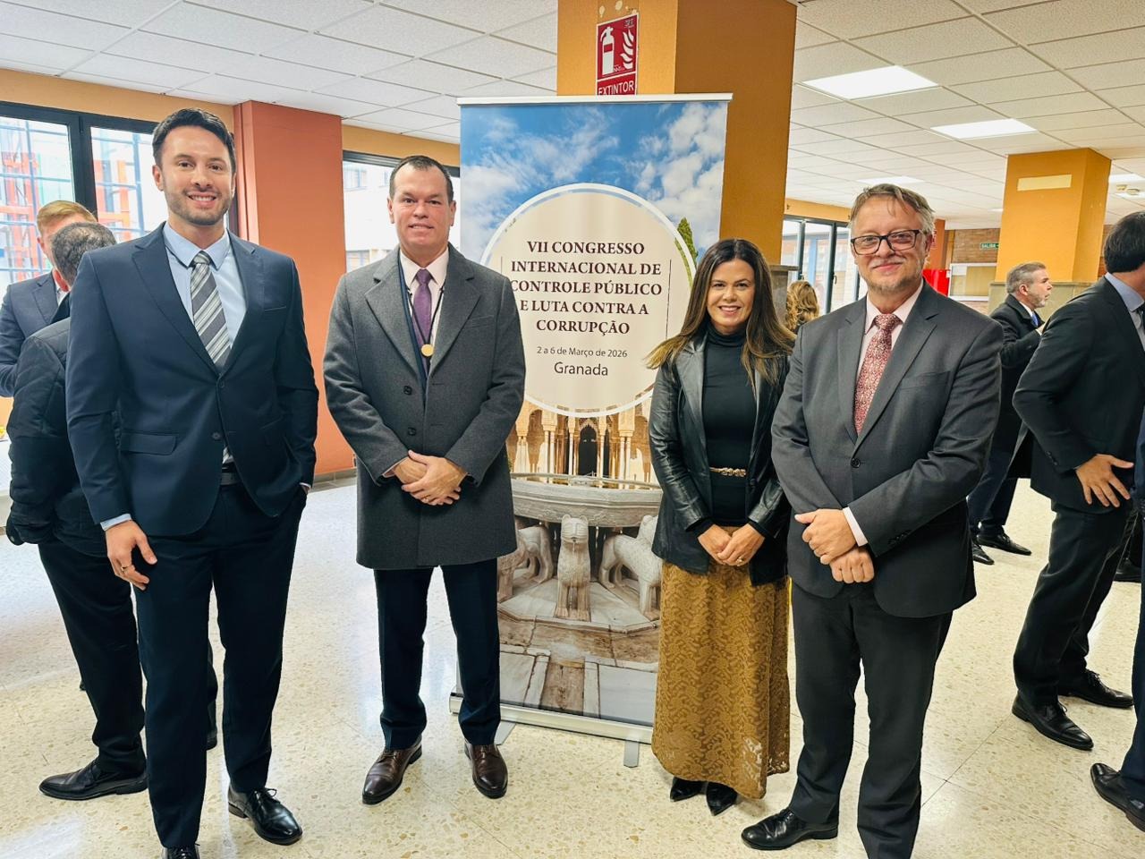 Foto horizontal que reúne o conselheiro Adircélio de Moraes Ferreira Júnior ao lado dos procuradores Cibelly Farias, Diogo Ringenberg e Leandro Ocaña Vieira. Eles estão em pé, lado a lado, em frente a banner que exibe o título do congresso internacional. Todos vestem trajes formais e sorriem. Ao fundo, há mais pessoas circulando.  