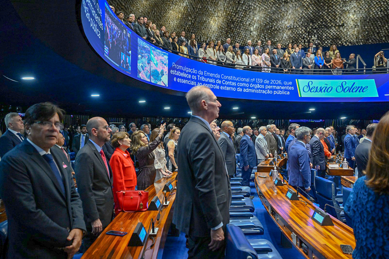 Foto horizontal de perfil do presidente do TCE/SC, conselheiro Herneus De Nadal, acompanhando a sessão solene de promulgação da EC. Ele é um homem branco, de cabelos claros. Está em pé, em meio a mais pessoas. Ao fundo, telão curvo mostra o título da sessão. 