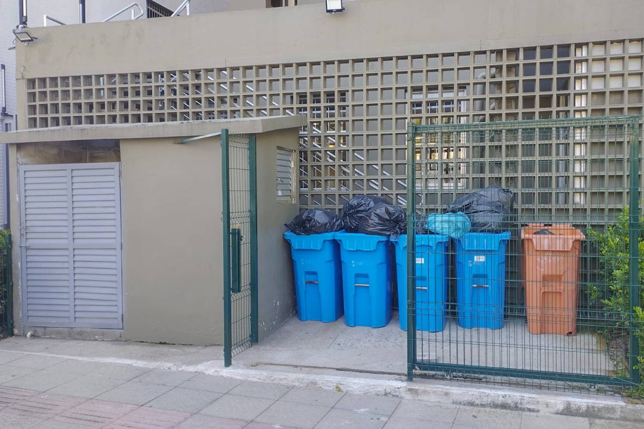 Foto horizontal da central de resíduos antes do PGRS. Há várias lixeiras cheias posicionadas no exterior do TCE.
