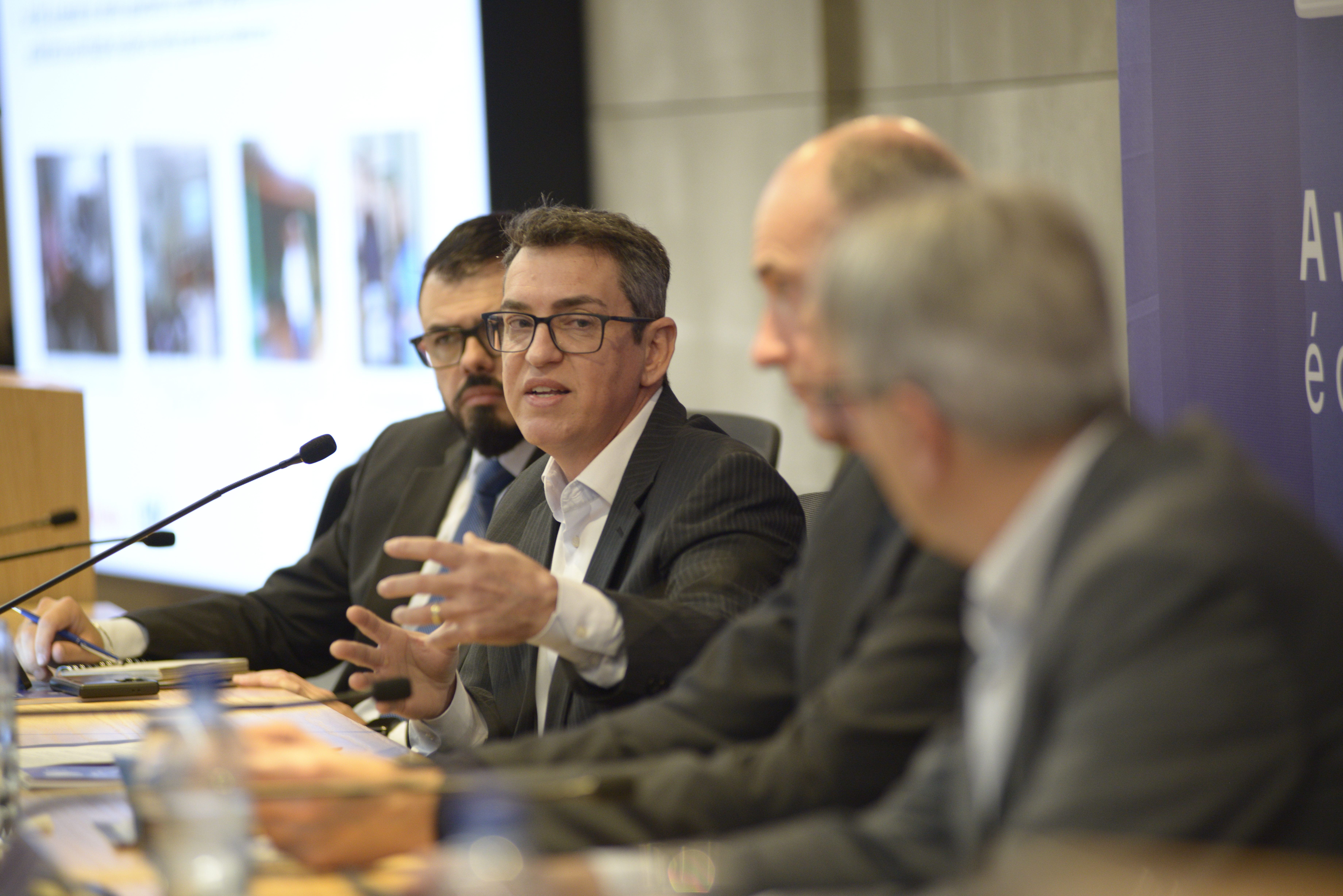 Foto horizontal do auditor fiscal de Controle Externo Hemerson Garcia falando aos demais. É um homem branco, de cabelos escuros. Usa óculos de grau e veste paletó. Está sentado em meio aos outros debatedores. 