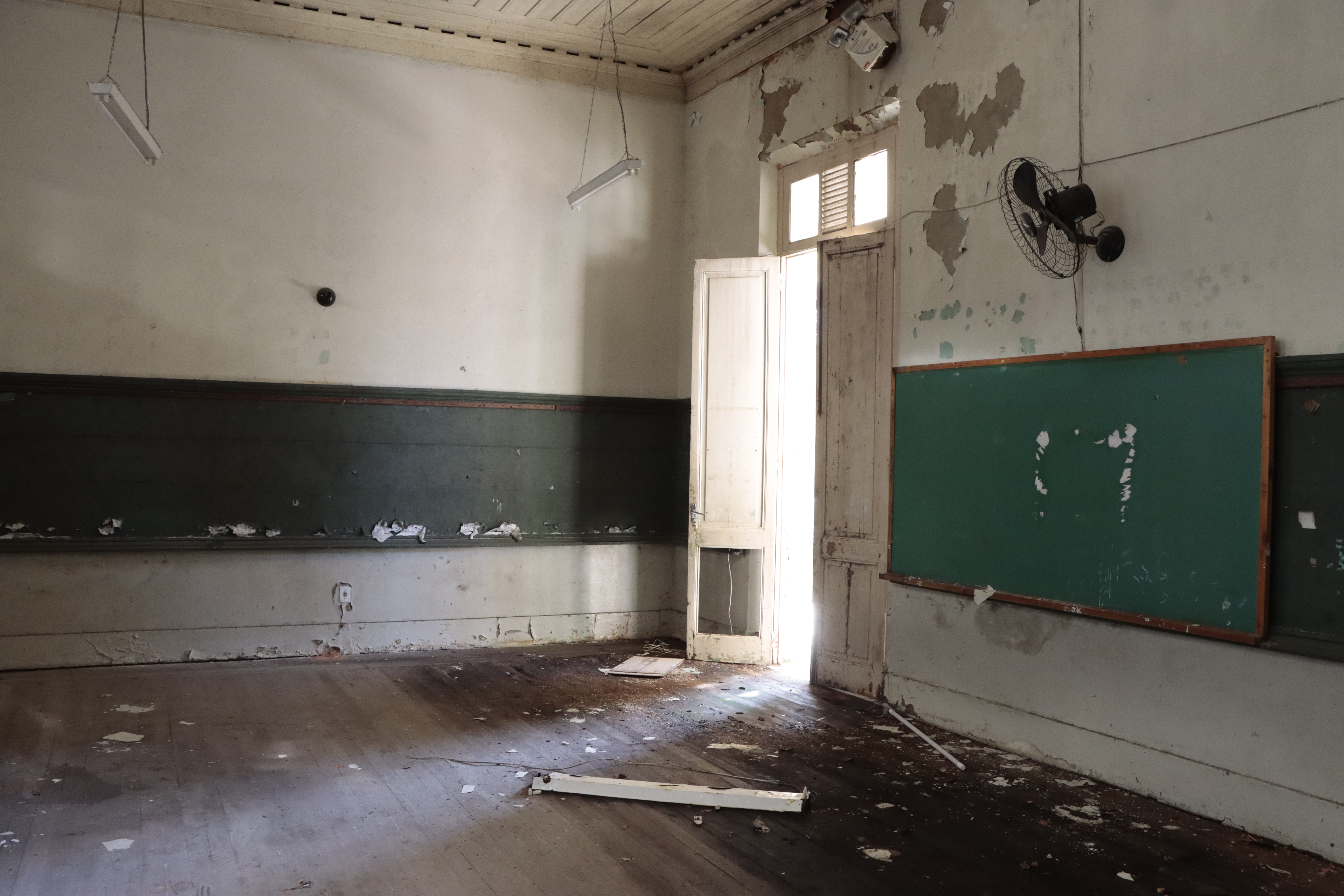 Foto horizontal do interior do prédio da Escola. O ambiente está  bastante degrado pelo tempo. Portas e janelas quebradas e paredes com infiltração.