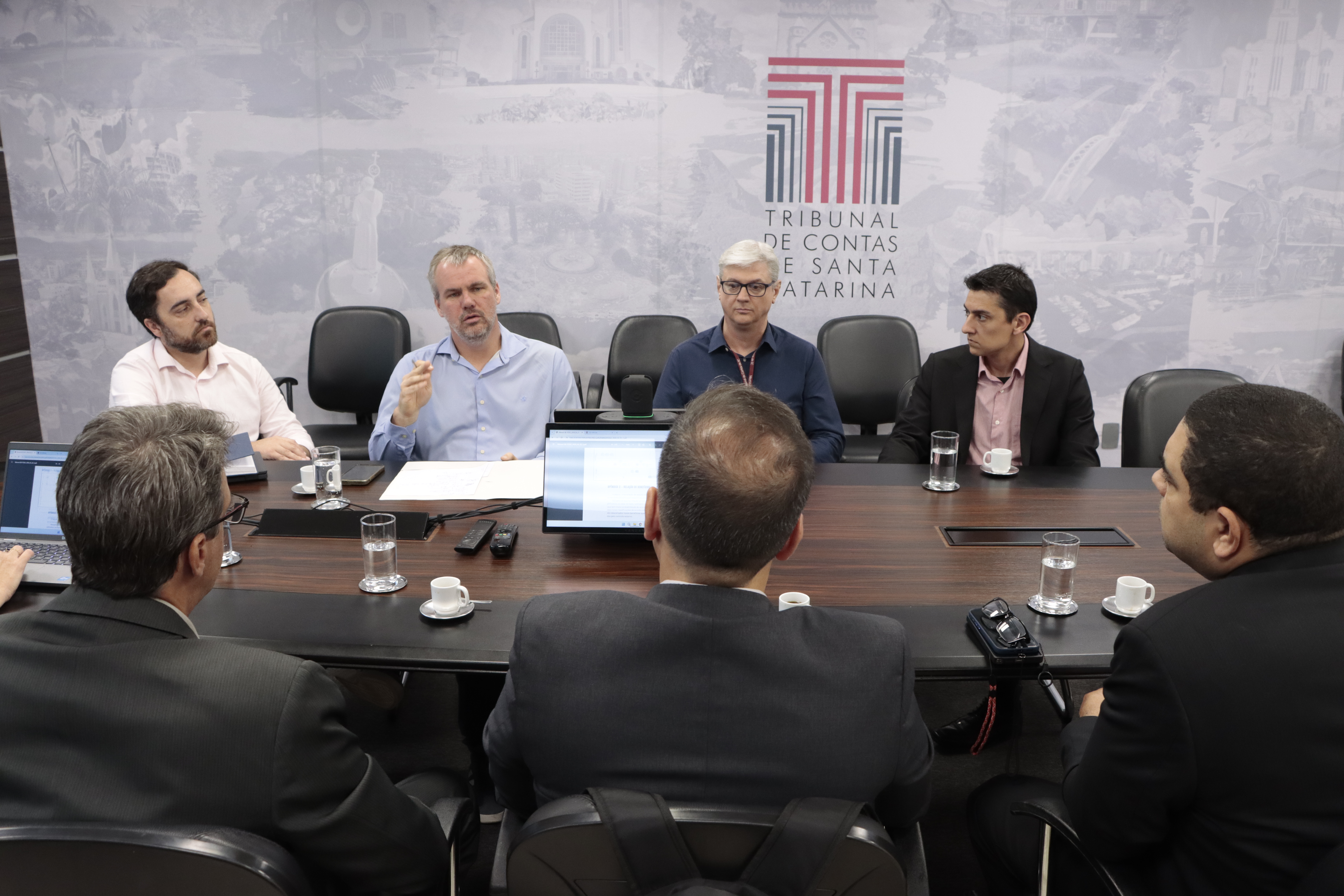 Foto que mostra sete homens sentados em volta de uma mesa. São representantes do TCE/SC e do TCE-RJ. Eles conversam em reunião. Ao fundo, na parede, plotagem do logo do TCE/SC. 