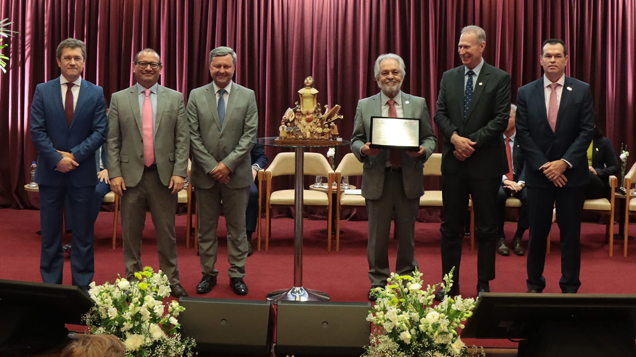 Foto horizontal com as autoridades em pé, lado a lado, no palco do auditório. Da esquerda para a direita, o conselheiro José Nei Ascari, o advogado Rodrigo Valgas, o advogado José Cristóvam, o homenageado, desembargador João Henrique Blasi, o presidente do TCE/SC, Herneus De Nadal, e o conselheiro Adircélio Ferreira Júnior. Eles posam para a foto. Blasi segura a placa recebida como homenagem e está ao lado da imagem do Boi de Mamão que recebeu de presente. O fundo é composto por cortinas bordôs.