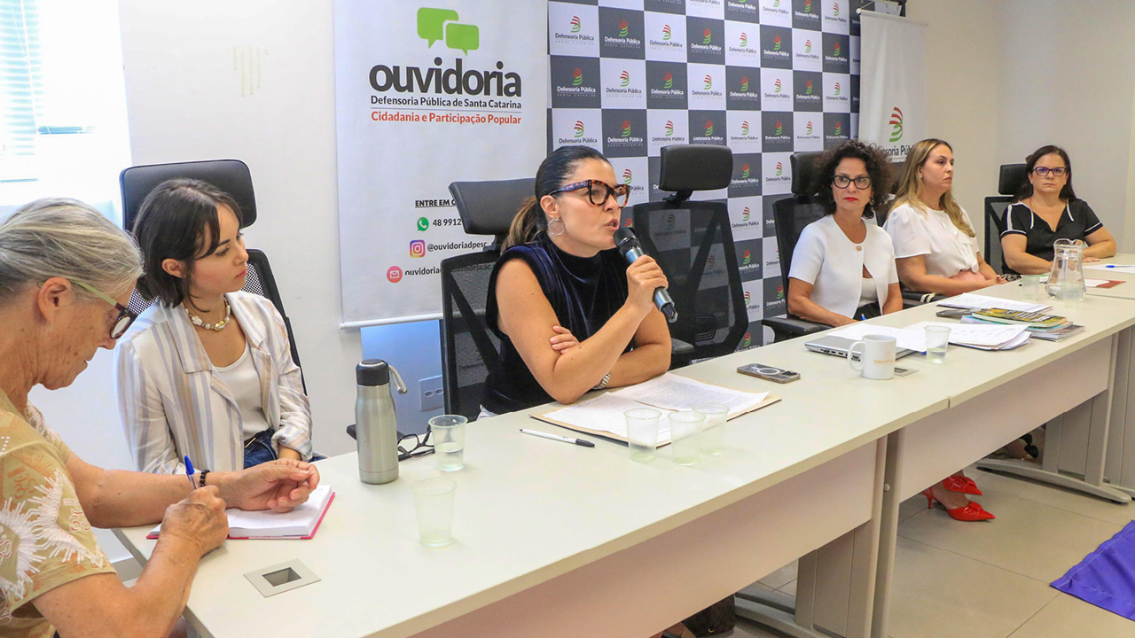 foto da mesa de debate. Ao microfone, a diretora adjunta Monique Portella , umamulher branca, de cabelos presos escuros. Ao lado dela, outras 5 mulheres compõem a mesa.