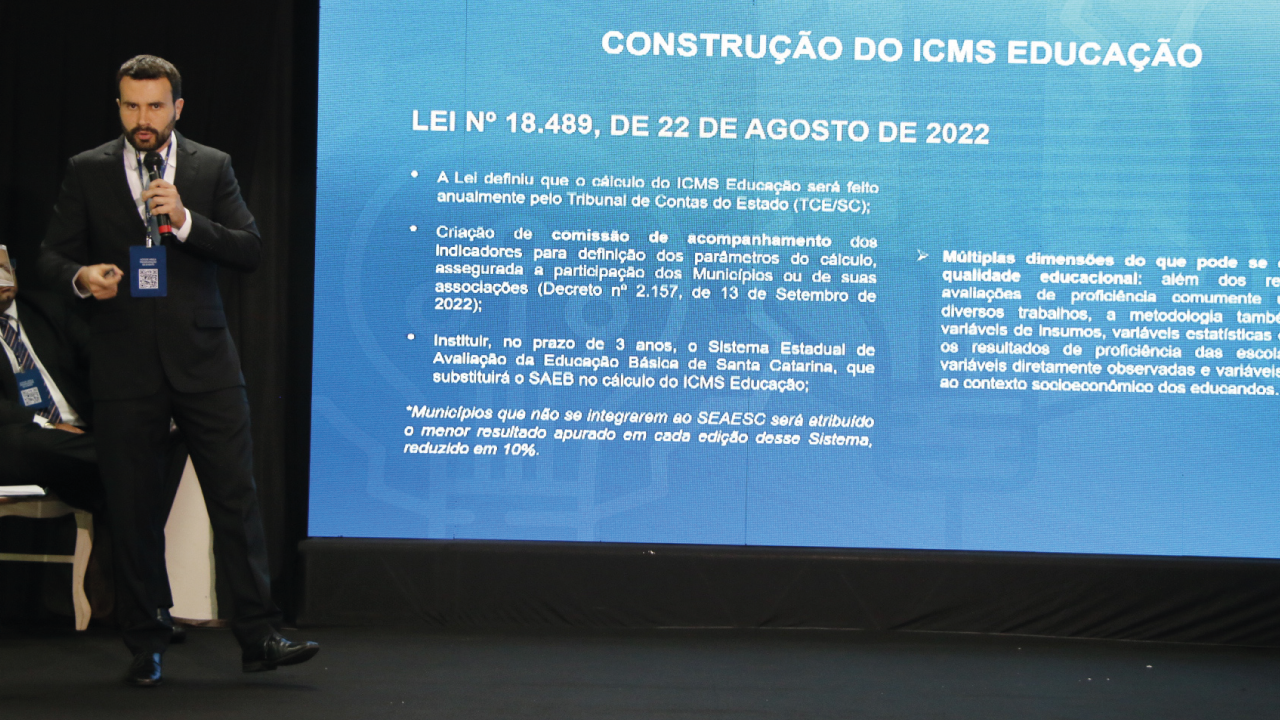 Foto do auditor fiscal de controle externo Silvio Sallum. Ele está em pé, no lado esquerdo da foto, ao lado do telão com trecho da apresentação.