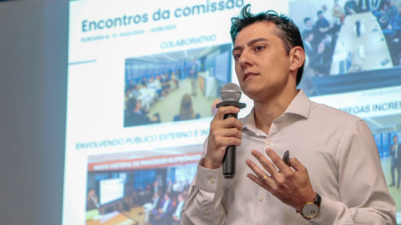 Foto horizontal do diretor da DTI. Ele está com o microfone na mão direita. Ao fundo dele, apresentação em slide projetada no telão..