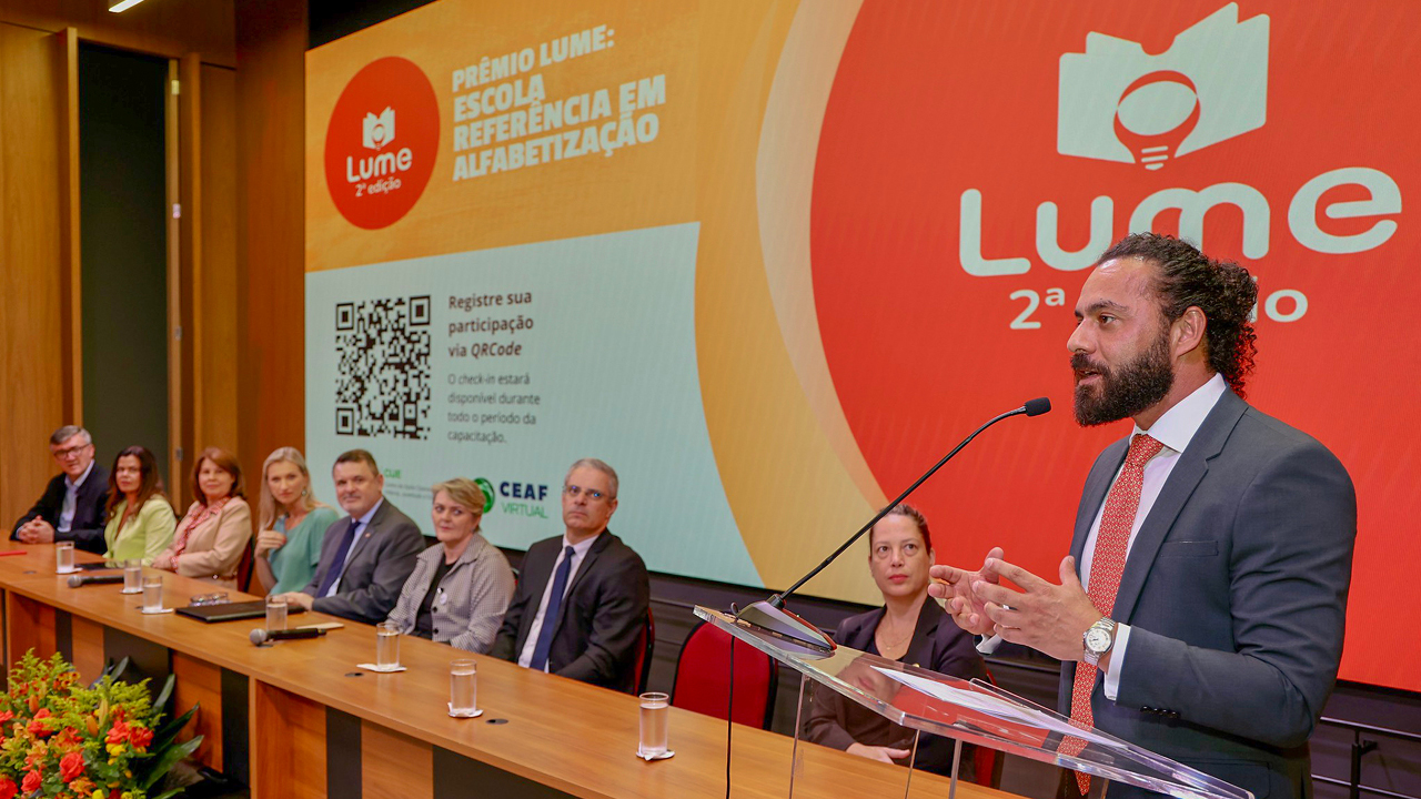 Foto do promotor de Justiça Eder Cristiano Viana falando ao púlpito no auditório do evento, Ele está em pé, à direita. À esquerda, há uma mesa de autoridades. Entre os presentes, o conselheiro substituto Gerson Sicca. Ao fundo, telão projeta logo do evento. 