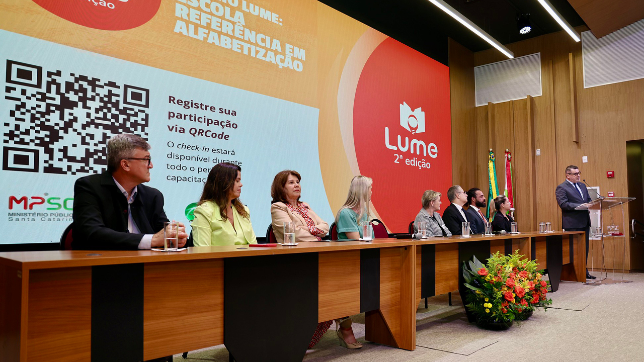 Foto da mesa de autoridades, no palco do auditório do evento. Entre os presentes, a procuradora Cibelly Farias e o conselheiro substituto Gerson Sicca. Ao fundo, no telão, é projetado o logo do evento. 