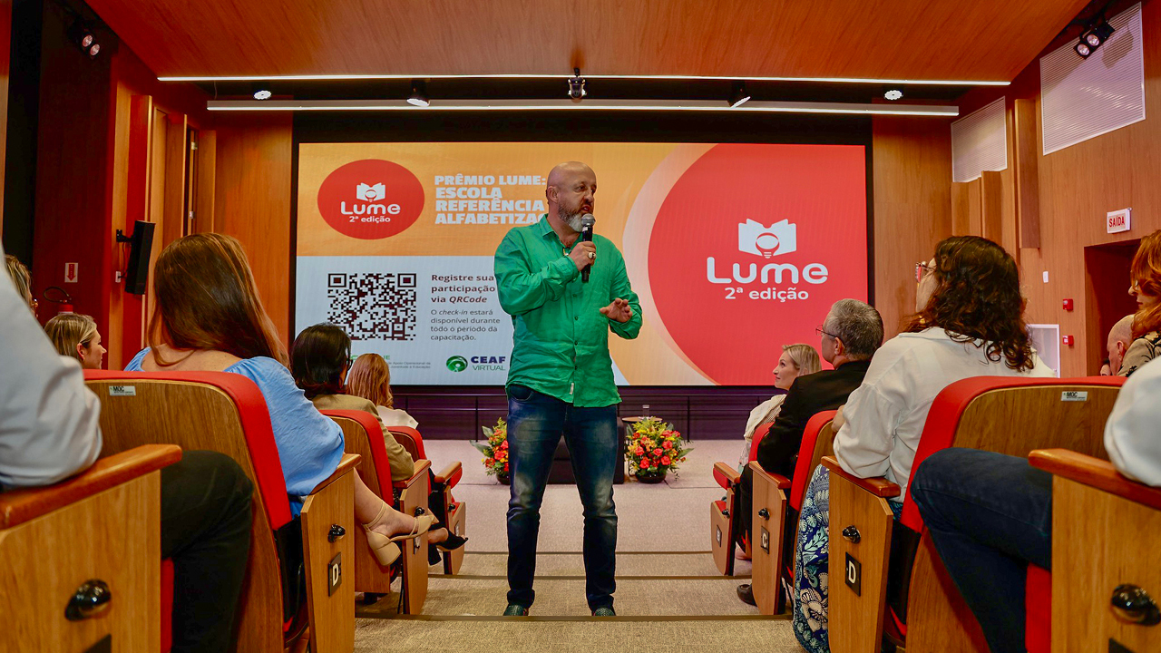 Foto do palestrante Fabrício Carpinejar falando ao público durante o evento do prêmio Lume. Ele está ao centro de um auditório, em pé, segurando o microfone nas mãos. Nas laterais, há pessoas sentadas em poltronas. Atrás dele, é projetado o logo do evento. 